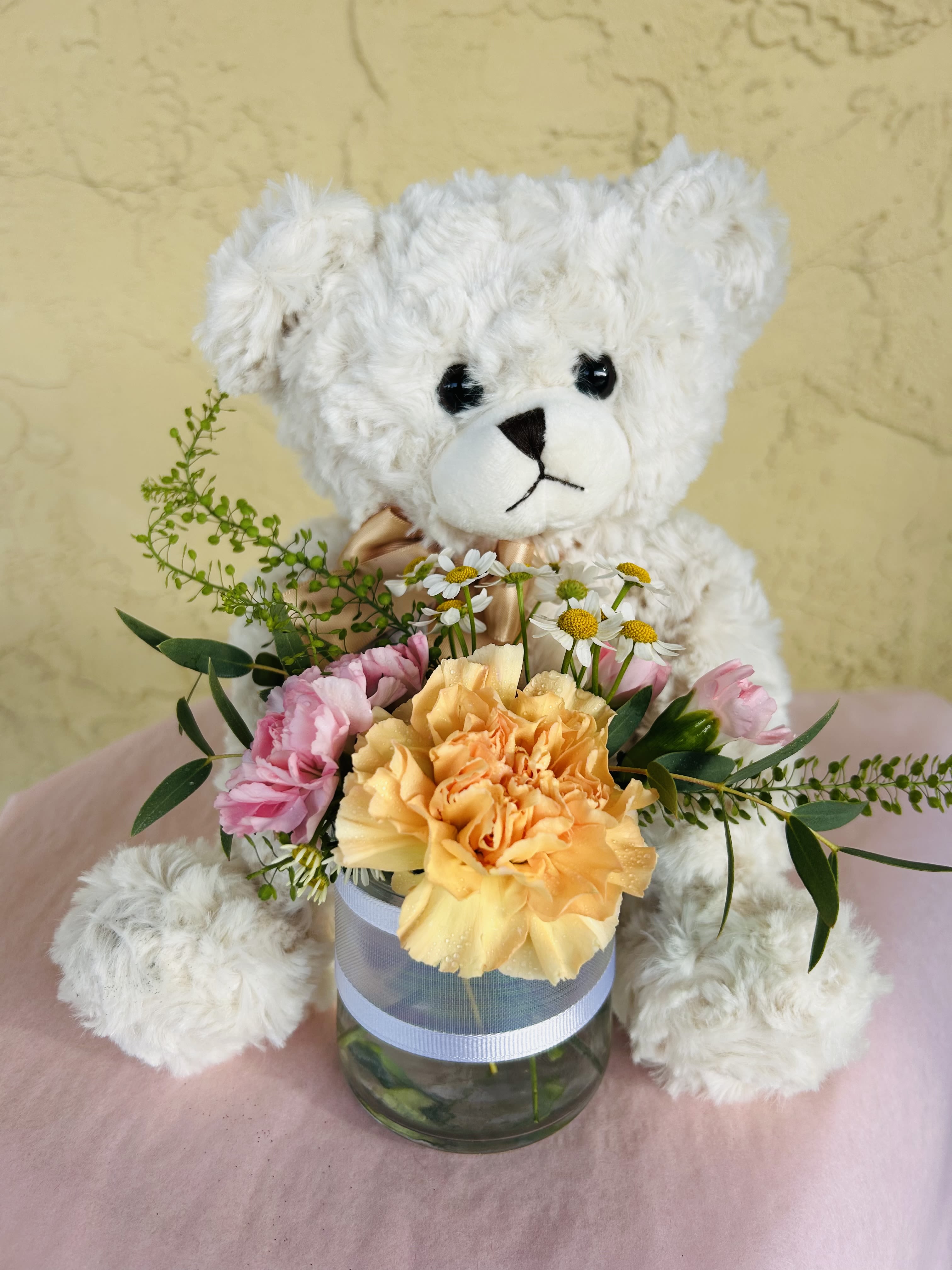 White teddy bear holding a small flower arrangement in a glass vase