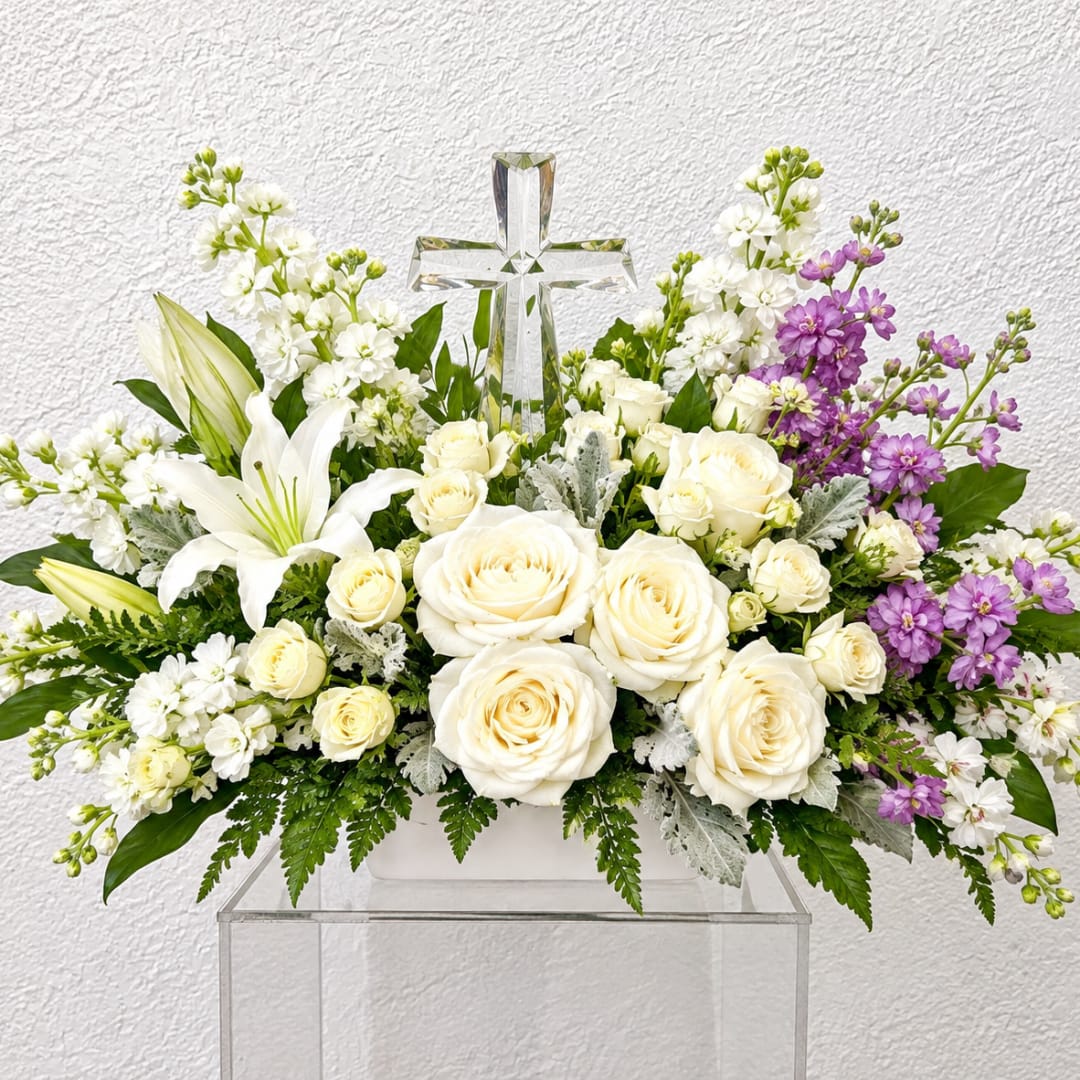 White roses and lilies arranged with purple flowers around a clear cross