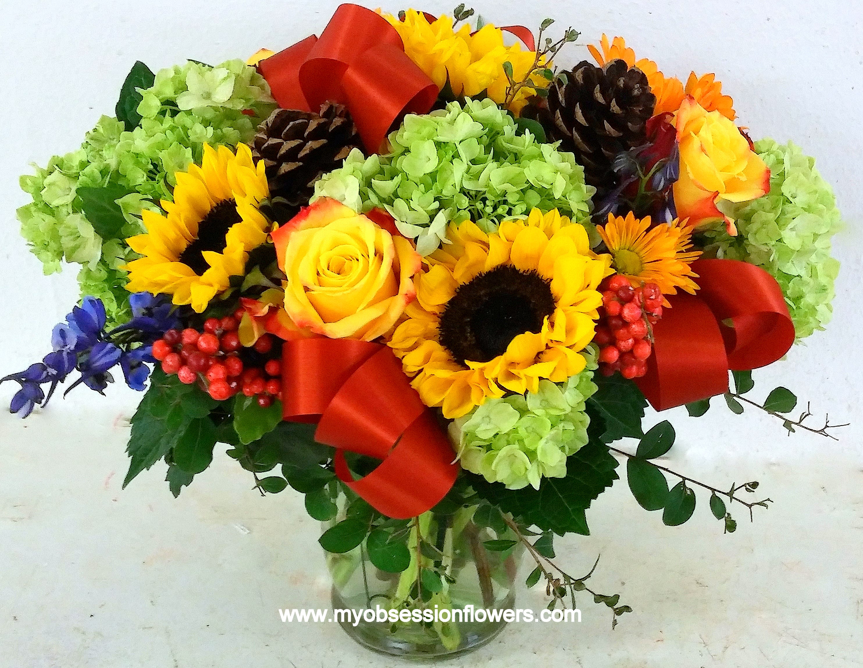 Bouquet of sunflowers, yellow roses, and green hydrangeas in a glass vase with red ribbon