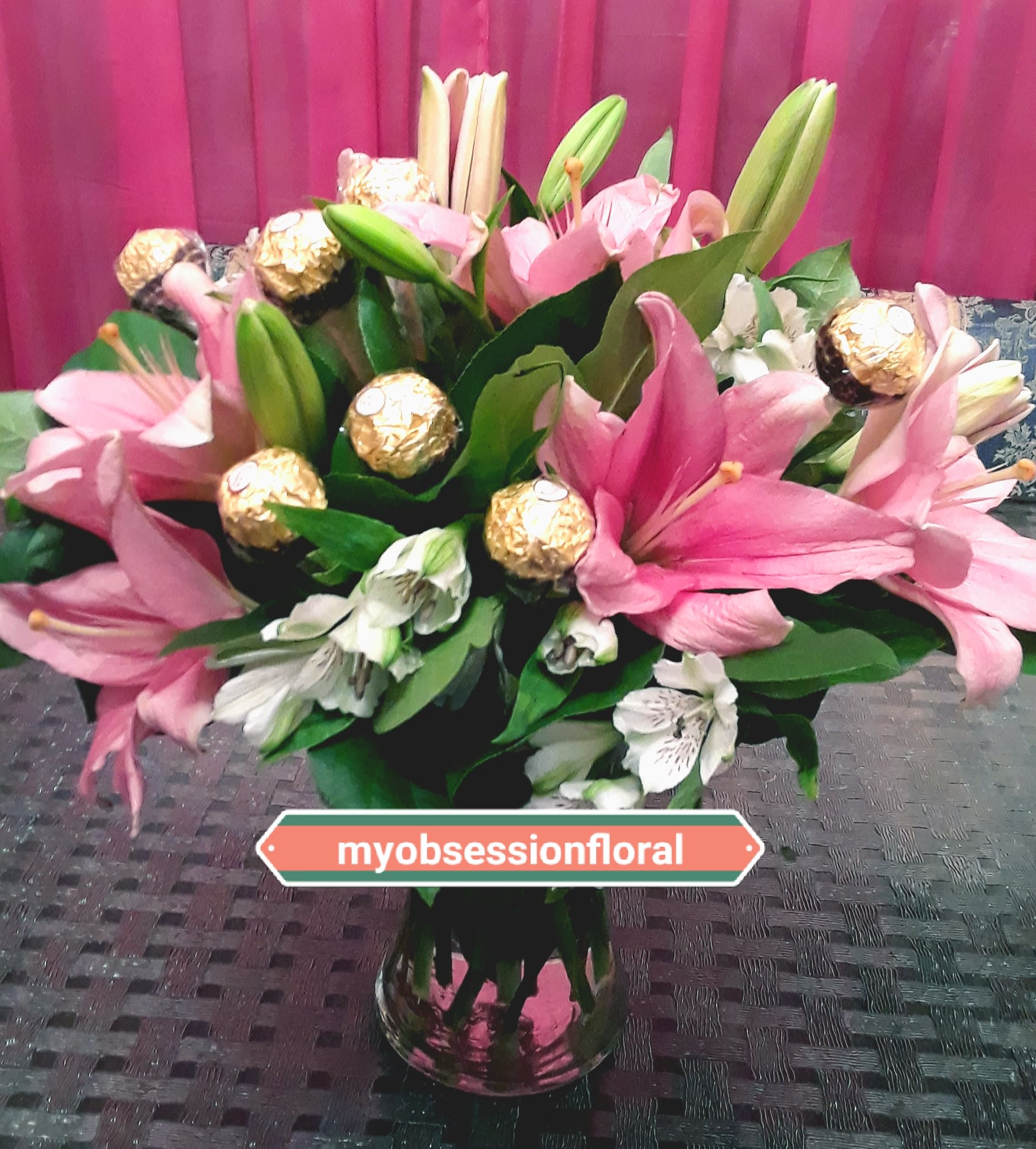 Pink lilies and white flowers arranged in a glass vase with gold-wrapped chocolates