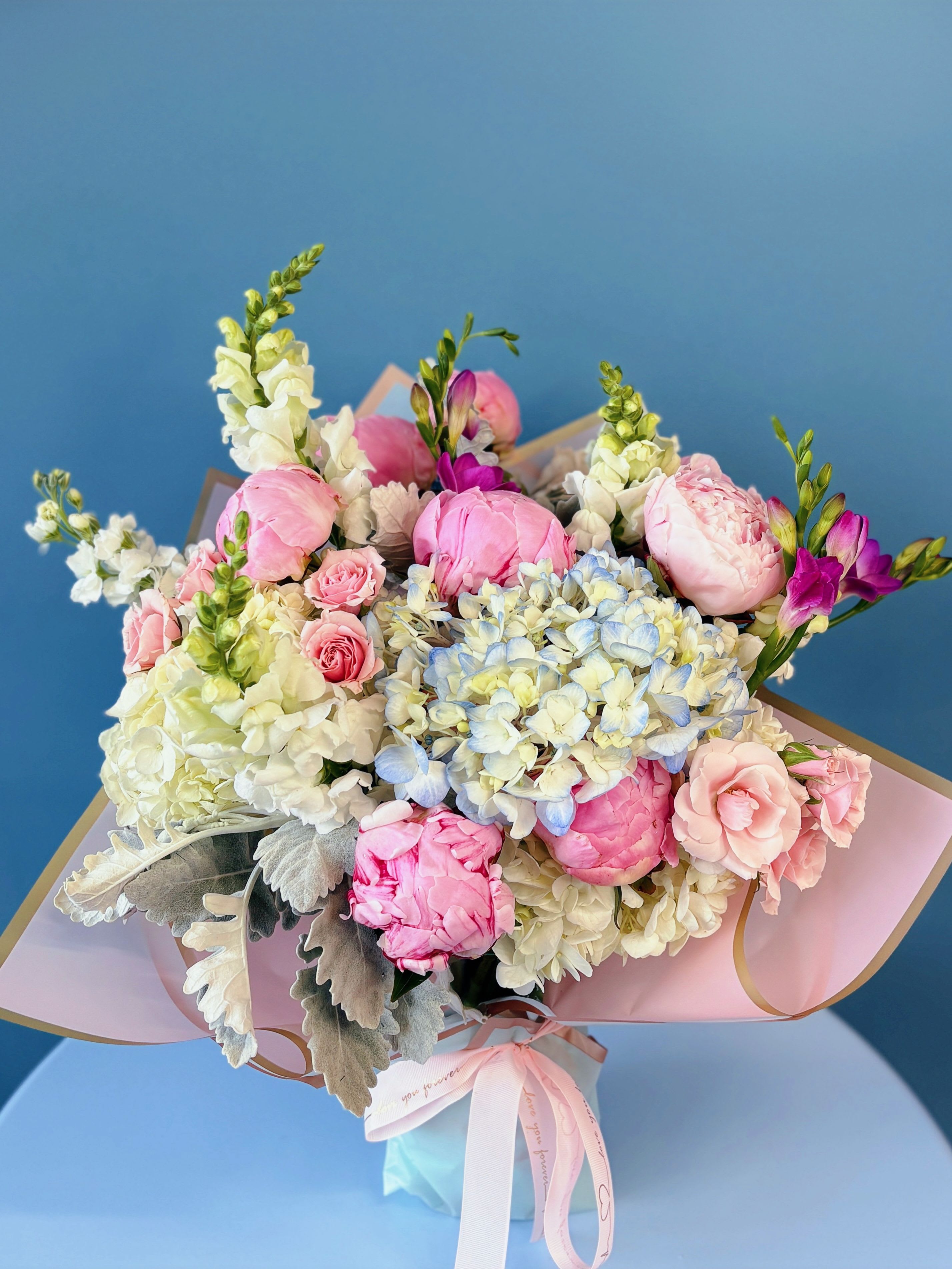 La Rose Grand Vase Bouquet - Experience the elegance of the La Rose Luxury Vase Bouquet, a stunning arrangement designed to elevate any space. Featuring Queen’s Crown roses, delicate ranunculus, lush hydrangeas, and airy pampas grass, this bouquet blends vibrant colors with soft, romantic textures.  Arranged in a designer's choice vase, this bouquet is perfect for celebrating special moments or simply brightening someone’s day. Its timeless beauty and luxurious details make it a centerpiece that speaks of artistry and love.  Luxury size: contains 18-20 premium blooms Grand Size:  contains 26-28 premium blooms