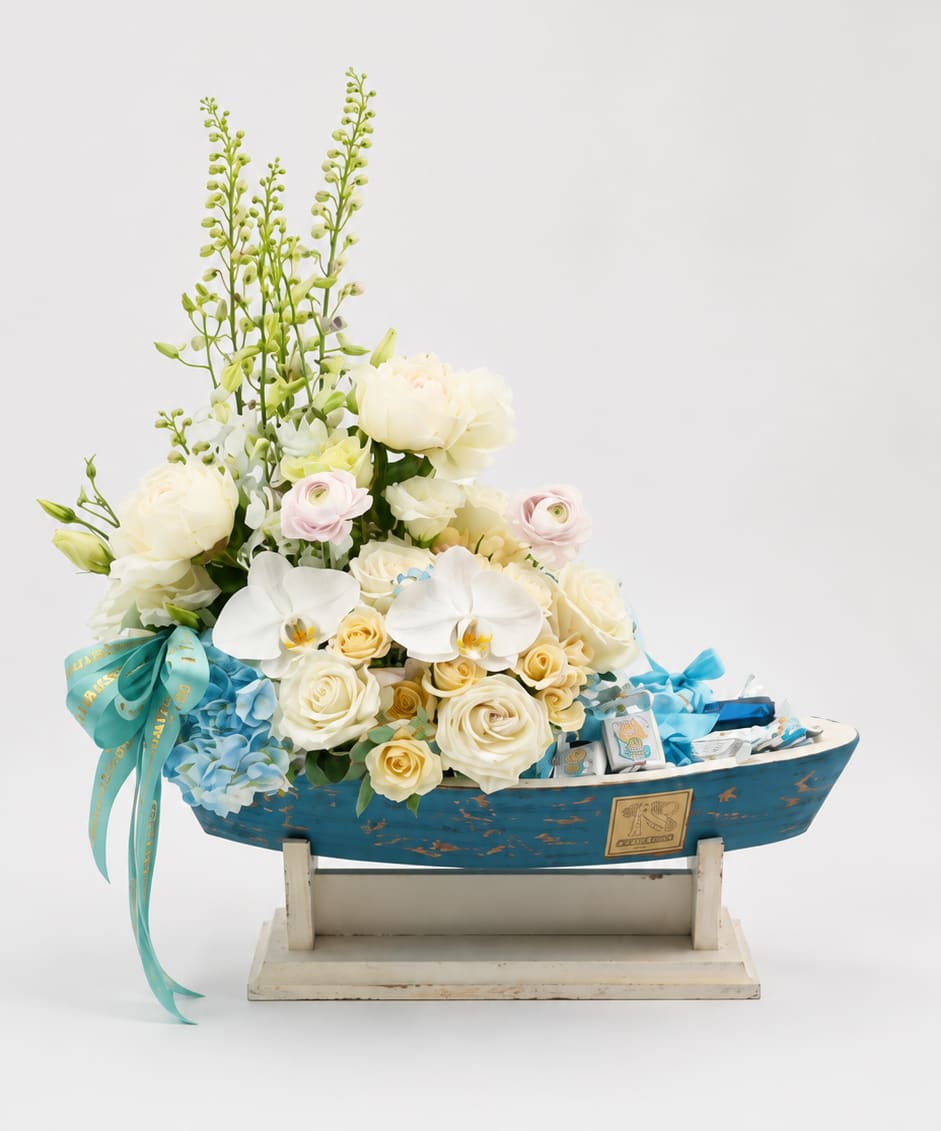 Baby Boy Chocolate Sailboat - A charming baby boy gift arrangement designed in a decorative blue boat container, filled with soft white and cream roses,  delicate seasonal blooms, and premium chocolates. Finished with a matching blue ribbon, this unique design is perfect for welcoming a newborn baby boy or celebrating a baby shower.