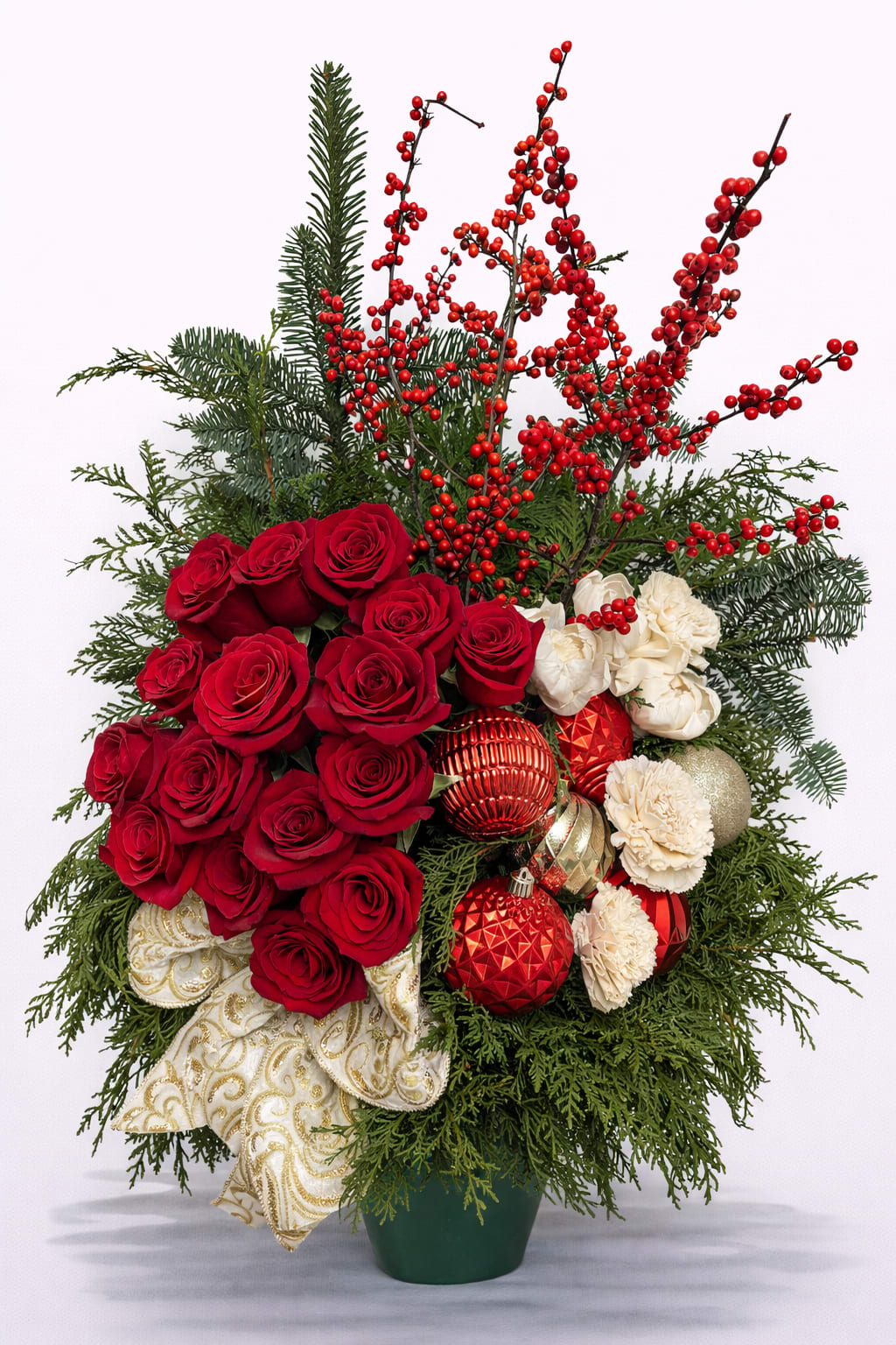Eternal Christmas Arrangement - Deep, velvety red roses and creamy white carnations create an immediate impression of warmth and affection in this holiday flower arrangement-perfect as a Christmas or winter centerpiece that will make the person you're thinking of feel cherished and celebrated. The Eternal Christmas Romance arrangement reads as classic and romantic: the rich red roses set a passionate tone while the white carnations add soft contrast, all conveying a message of love and festive cheer for holiday gatherings or a special winter surprise.  Lush evergreen branches form a textured backdrop, with sprays of bright red berries that add height and a natural, woodland feel. Shiny red and gold Christmas ball ornaments are tucked throughout for sparkle, and an ornate white-and-gold patterned ribbon introduces elegant decorative detail. The design is arranged in a simple green plastic container that suits seasonal décor and makes placement easy on a table or mantel. Sized to command attention without overwhelming a room, this arrangement brings traditional red, white, and green holiday color right into their home in Newington, CT.  This piece works beautifully as a holiday gift, a winter anniversary surprise, or a centerpiece for seasonal entertaining-she'll feel truly seen and celebrated when it arrives. Light care: keep in a cool spot away from direct heat, refresh water as needed, and enjoy the flowers for several days; order with same-day delivery where available for last-minute winter giving.