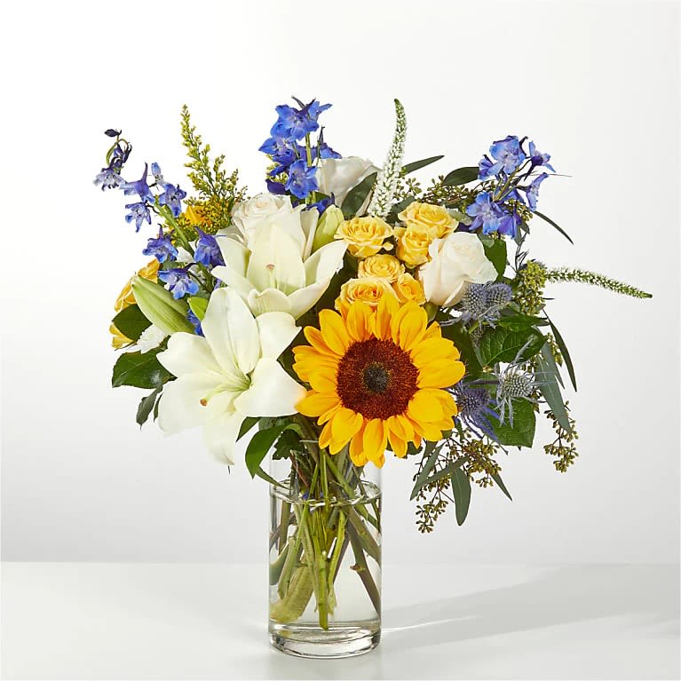 Sunny Bouquet - Bright, garden-fresh and instantly uplifting, this sunny flower arrangement pairs a bold yellow sunflower and soft yellow roses with large white lilies and sky-blue delphinium for a joyful birthday bouquet or cheerful "just because" gift. The mix of warm yellows, crisp whites, and cool blue accents reads lively yet balanced; it's the kind of arrangement that makes the person you're thinking of feel genuinely celebrated and noticed.  Arranged in a clear cylindrical glass vase, stems are visible for a fresh, natural look-flowers extend outward in a slightly airy, full composition that feels hand-gathered. The design also includes white daisies and greenery that add texture and movement among the focal blooms. Available in three sizes (Standard, Deluxe, Premium), larger sizes offer a noticeably fuller, more expansive presentation while all versions maintain the same bright, layered palette.  Perfect for birthdays, congratulations, thank-you gifts, or simply brightening someone's day, this bouquet arrives ready to display in the included glass vase. For best enjoyment, keep in fresh water, out of direct sunlight, and recut stems every few days to extend the life of the lilies, roses, sunflowers, delphinium, and daisies.