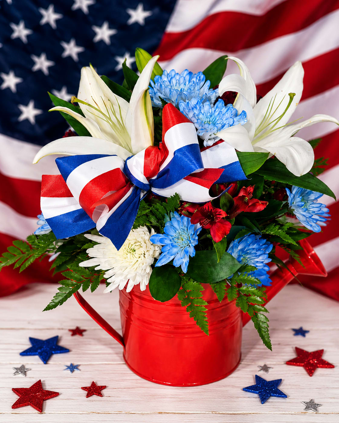 All American Watering Can - Celebrate the spirit of America with this stunning red, white, and blue floral arrangement! Designed in a charming red watering can, this festive bouquet features elegant white lilies, vibrant blue chrysanthemums, rich red blooms, and lush greenery accented with a patriotic ribbon bow. Perfect for Memorial Day, Fourth of July, Veterans Day, or showing your American pride, this eye-catching centerpiece brings beauty and celebration to any home or event. ️