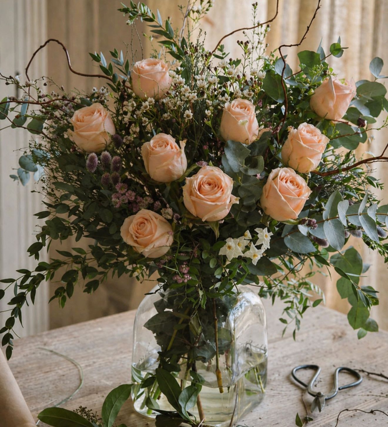 Peachful Chamber - A soft, romantic composition that feels like a quiet moment in bloom. Peachful Chamber features lush peach roses nestled among airy greenery and delicate touches of wax flower, creating a naturally graceful silhouette. The subtle filler adds a light, whimsical texture, while the roses bring warmth and elegance to the arrangement.  Designed with an organic, garden-inspired style, this piece offers a gentle balance of softness and movement—perfect for bringing calm beauty into any space.