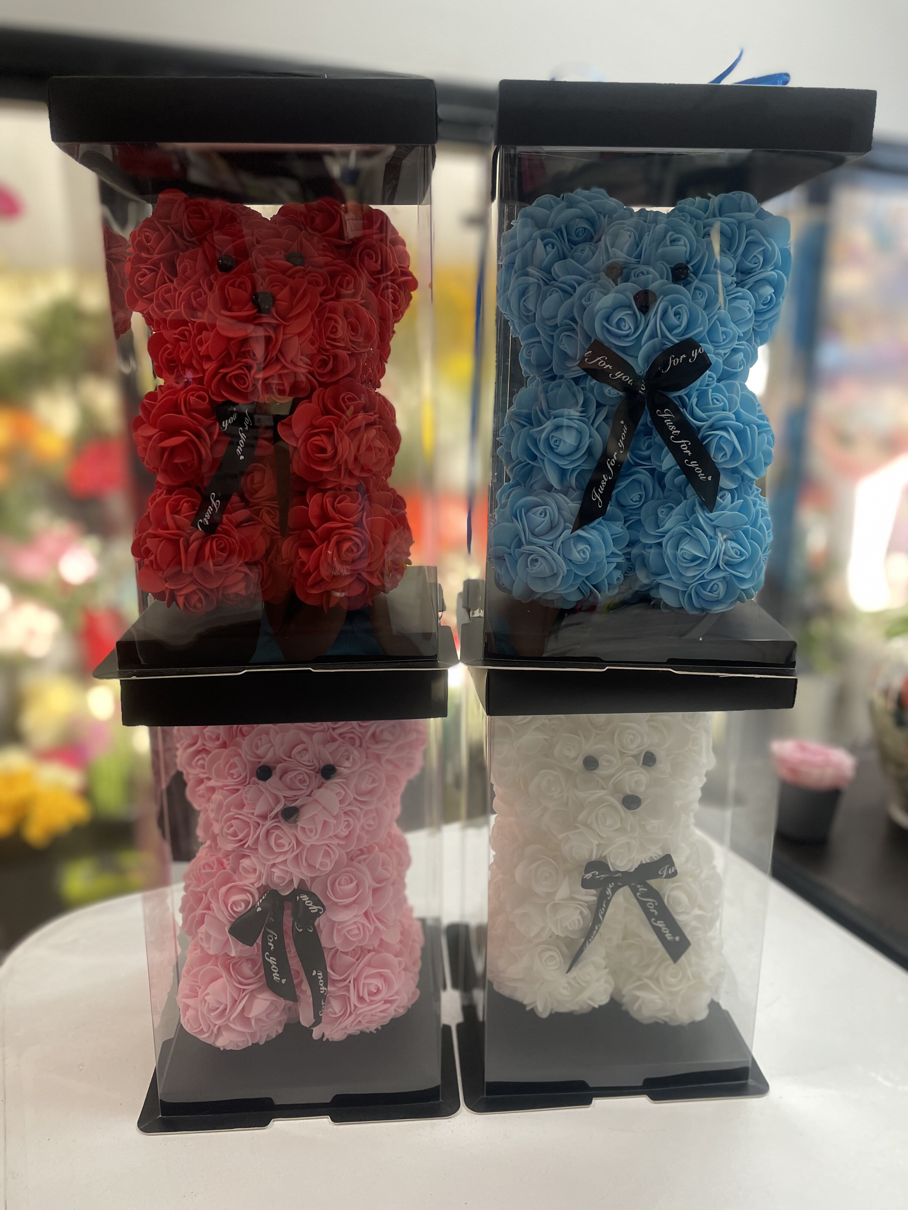 Four rose teddy bears in clear display boxes, in red, blue, pink, and white.