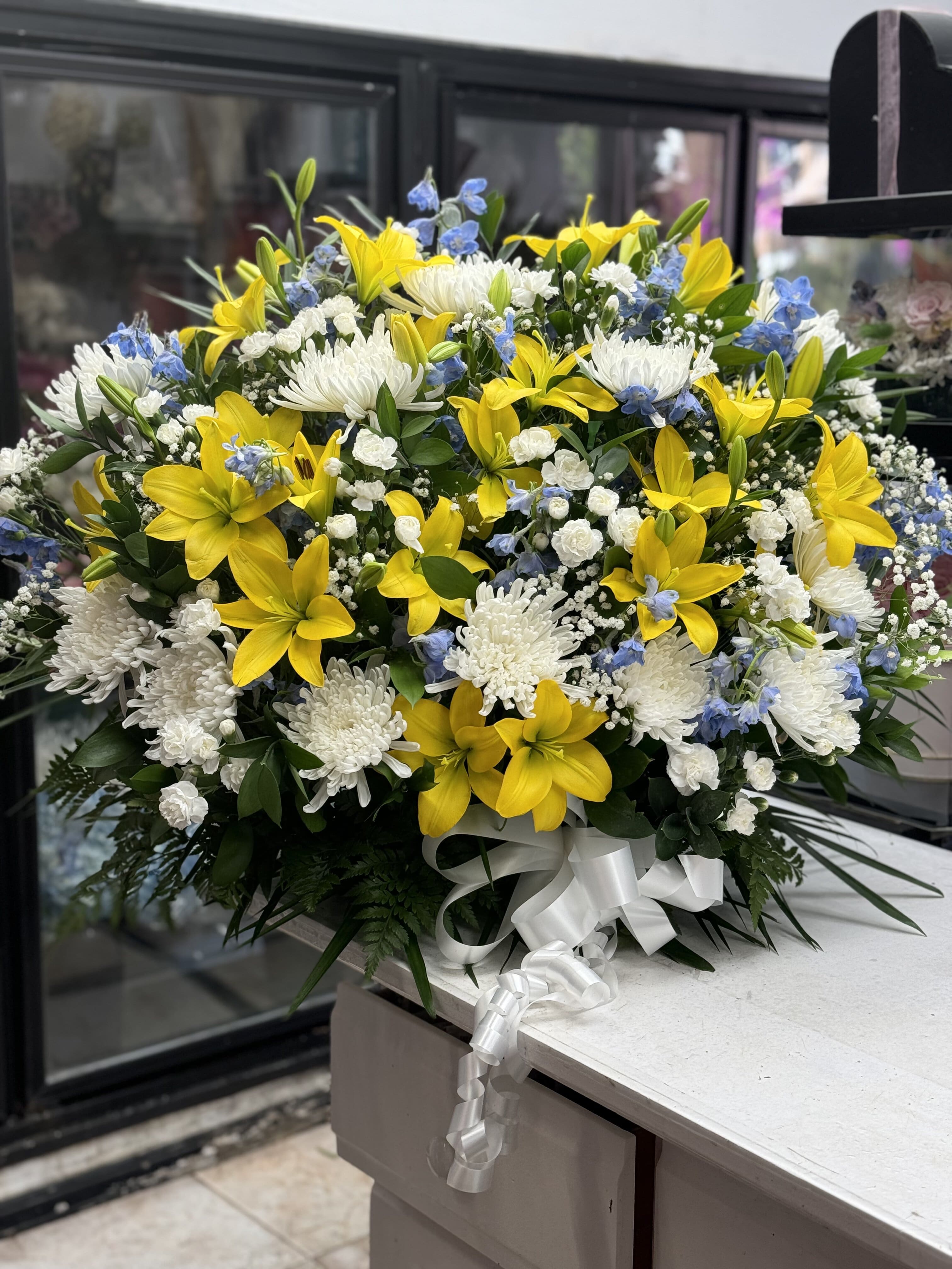 Large bouquet of yellow lilies, white chrysanthemums, and blue accents with a white ribbon