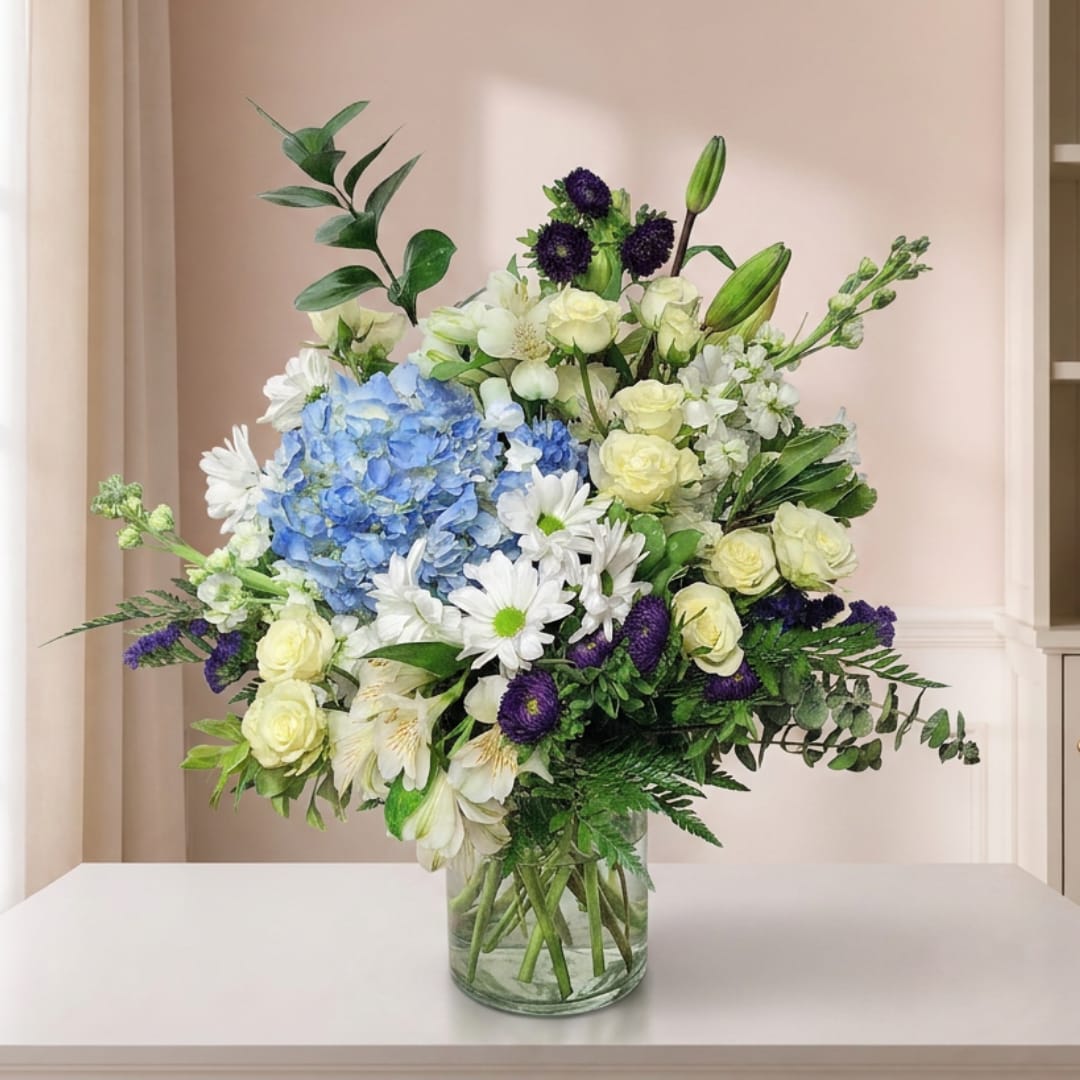 Mixed bouquet of blue hydrangeas, white daisies, and pale roses in a glass vase