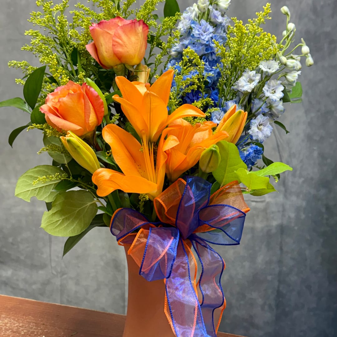 Orange roses and lilies in a tall vase with blue flowers and a ribbon bow