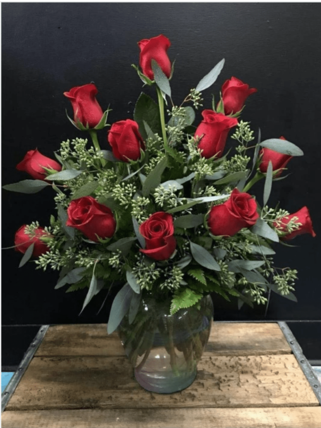 Tanzania Red Rose Arrangement in Clear Glass Vase - A dozen red roses gathered into a rounded, full design create an immediate classic statement-this romantic flower arrangement is perfect as an anniversary bouquet, a Valentine's Day gift, or any moment when you want the person you're thinking of to feel deeply loved. The rich red blooms read as timeless devotion while the clear glass vase keeps the look polished and formal, so she'll feel truly celebrated the moment these arrive.  The arrangement pairs a dozen or so long-stem roses with mixed greenery and delicate filler sprigs for a lush, layered texture and strong red-and-green contrast. The compact, rounded shape sits neatly in a clear glass vase that showcases the stems and keeps the focus on the velvety red petals and fresh foliage. Size and stem length are arranged to create a balanced, table-friendly display that suits both intimate dinners and larger room placement.  Send this when you want to mark a romantic milestone or simply remind someone they're cherished-it's the kind of bouquet that reads as thoughtful and elegant without fuss. Care is simple: keep the vase filled with fresh water, trim stems if needed, and refresh water every few days to extend bloom life.