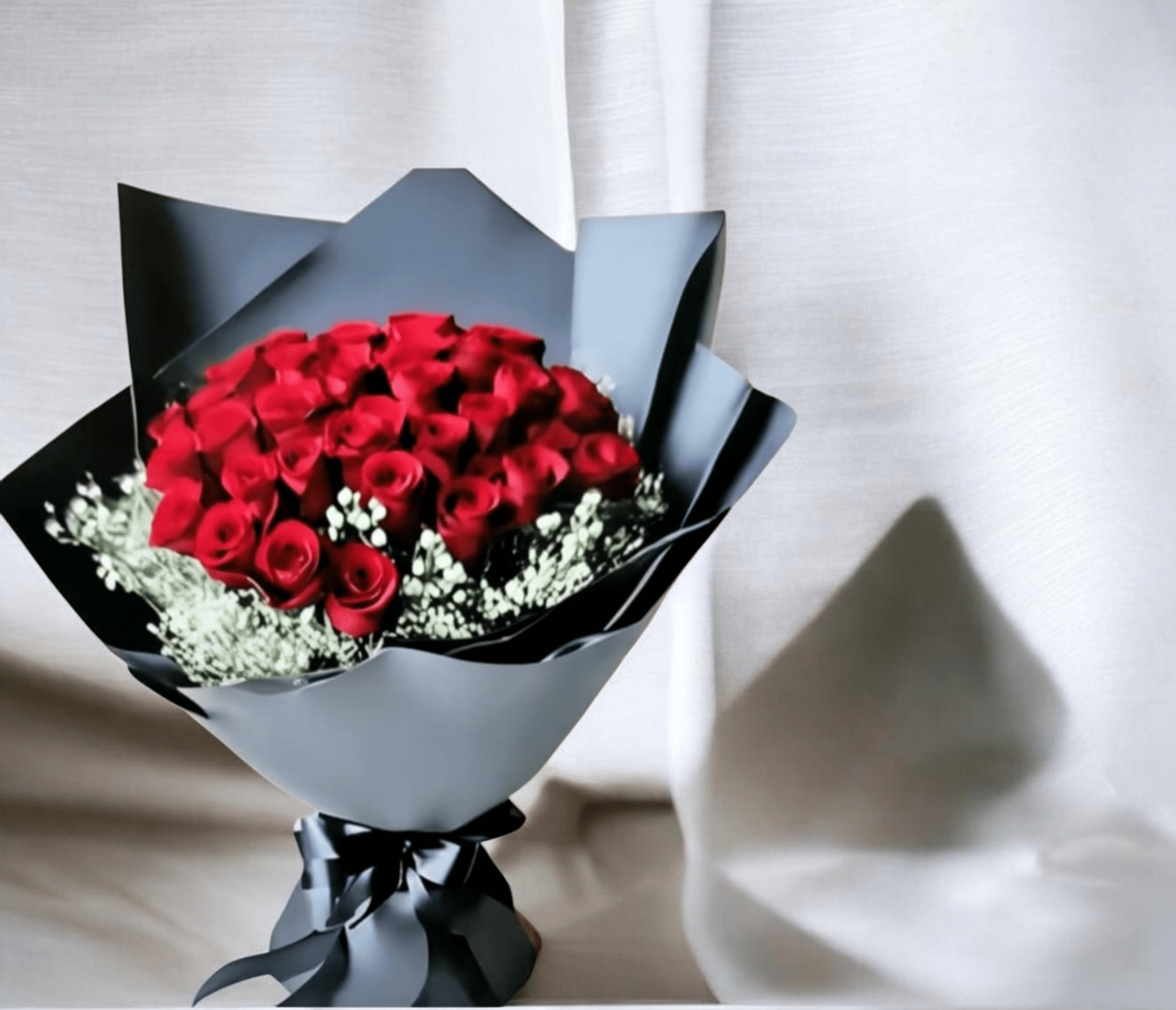 The Queen Red Rose Bouquet - A dense, rounded cluster of velvety red roses makes an immediate statement-THE QUEEN is a romantic bouquet perfect for an anniversary, Valentine's Day, or any moment you want the person you're thinking of to feel deeply adored. The dramatic contrast of rich red blooms against layered black and gray decorative paper gives the arrangement a polished, formal look that reads both luxurious and intimate, sending a clear message of passion and devotion.  Carefully hand-tied, the compact bouquet is finished with dark ribbon and trimmed with small white filler flowers that add soft texture and delicate highlights around the edges. Decorative wrapping paper and paper layers create a striking silhouette that holds its shape from presentation to delivery. This arrangement arrives ready to gift; recommend keeping the roses in fresh water, away from direct heat, to extend their beauty for as long as possible.