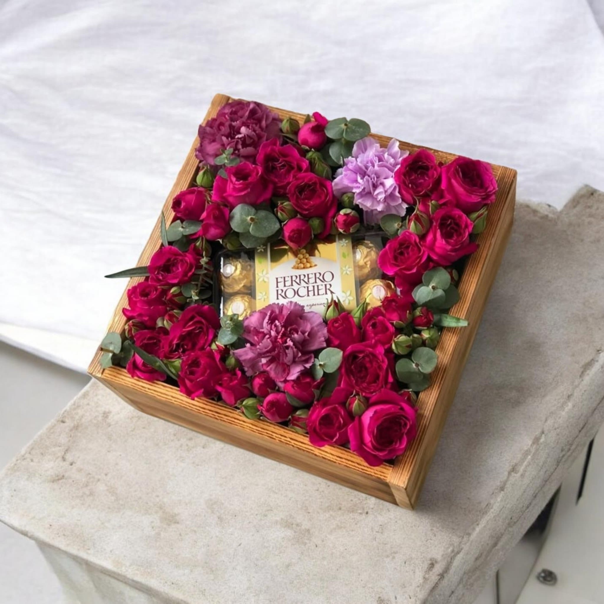 Virginia Mix Rose and Carnation Box Arrangement with Chocolates - Bright magenta roses take center stage in this romantic flower arrangement, making it an ideal birthday bouquet, anniversary gift, or a sweet way to say "thinking of you." Framed around a central compartment of Ferrero Rocher chocolates, the flower arrangement and treats combine to create an unforgettable gift that feels polished and indulgent for someone you love.  The arrangement is presented in a square wooden gift box filled with vibrant pink roses, purple carnations for contrast and texture, white accents, and small round eucalyptus-like greenery that creates a lush, compact border. The low, box-style design sits neatly on tabletops and mantels, delivering both visual impact and easy display.  Perfect for the person who enjoys classic blooms with a decadent twist, this keepsake wooden box makes the moment feel thoughtfully curated. Size and care: box-style arrangements are low-maintenance-keep flowers in a cool spot away from direct sunlight and refill water as needed for the freshest look; chocolates come ready-to-enjoy.