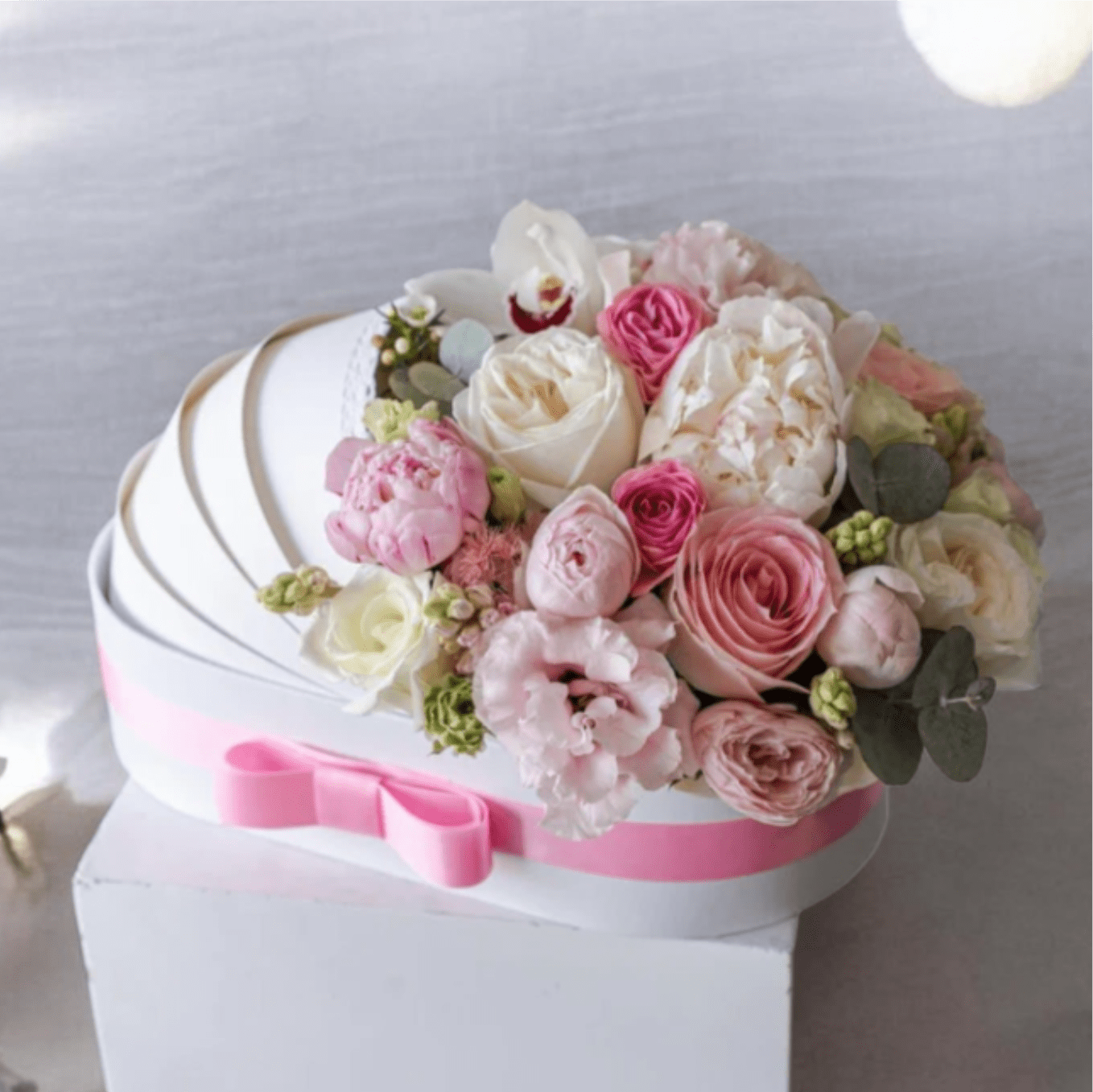 Welcome Baby Girl Pink and White Hat Box Arrangement - A lush cluster of pink and white roses, ranunculus, lisianthus and delicate orchid accents arranged in a heart-shaped gift box or a round white hat box makes a tender welcome for a newborn or a joyful congratulations for new parents. This welcome baby girl arrangement reads soft and celebratory-the blush roses and creamy whites create a gentle, feminine palette while the muted green accents add freshness that keeps the bouquet feeling light and modern.  Each option is presented in a polished white container: a heart-shaped box topped with a satin pink ribbon or a white pedestal/hat box tied with the same pink ribbon, both filled densely so the blooms form a full, rounded top. Textures range from the layered petals of ranunculus and roses to the silky orchids and airy lisianthus, giving visual depth and a luxurious feel without being fussy.  Send this as a sweet way to celebrate a newborn daughter, a baby shower gift, or a thoughtful hospital visit-she'll feel warmly seen and celebrated. Boxes arrive ready to display on a bedside table or nursery dresser; to keep blooms fresh, place in a cool spot away from direct sun and follow any water-care card included with the arrangement.
