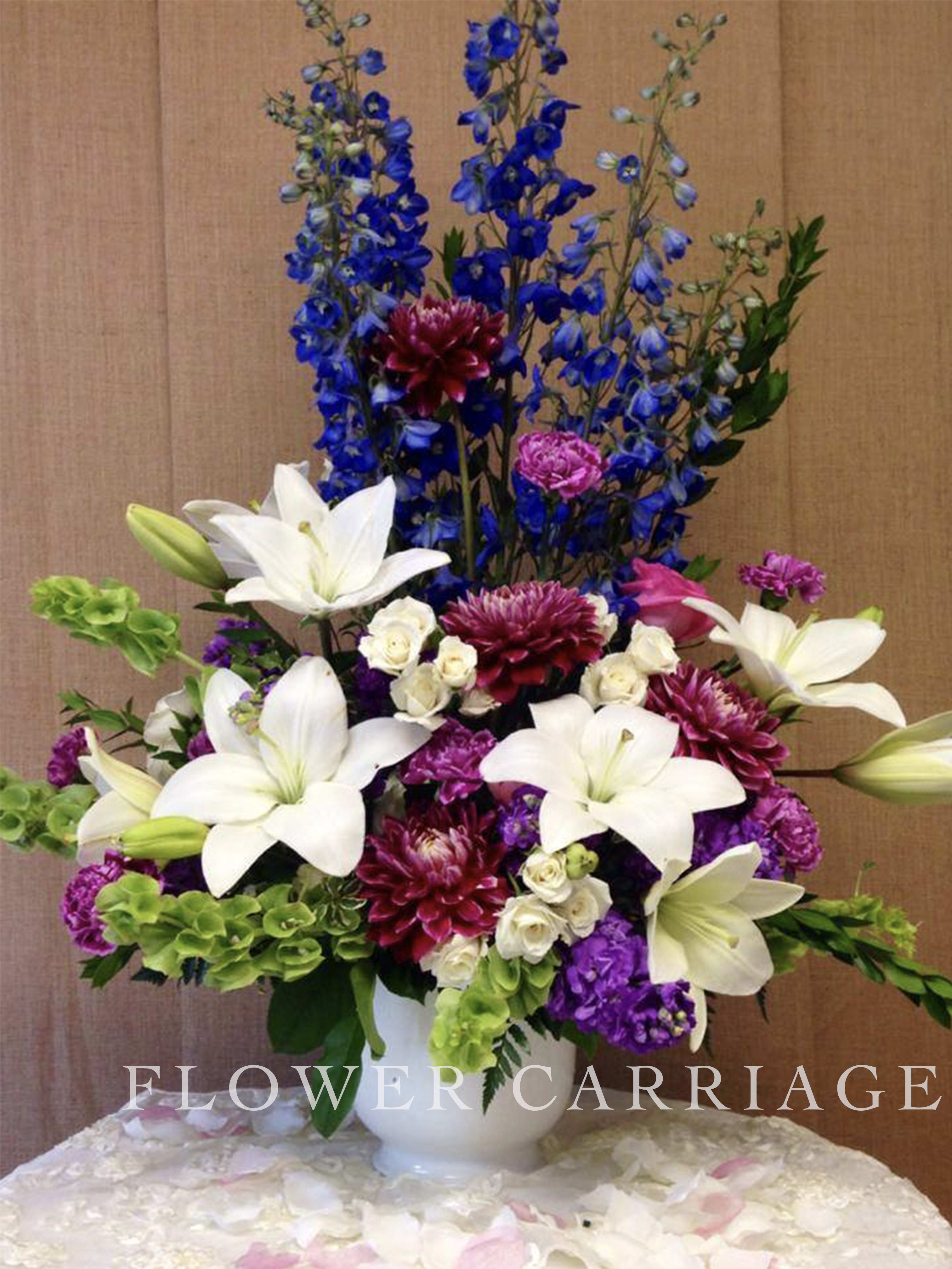 Tall bouquet of white lilies, blue delphinium, and purple blooms in a white vase