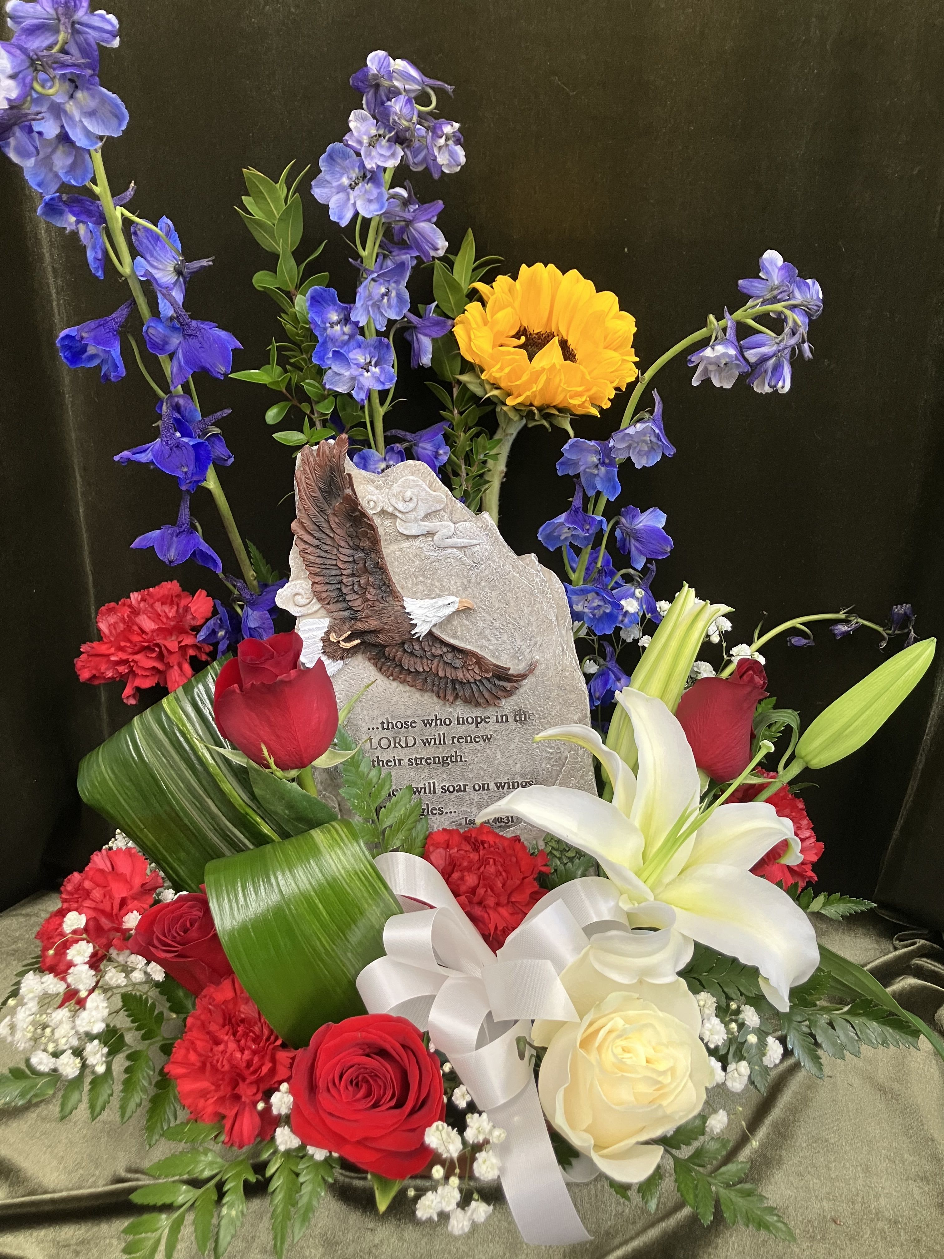 Wings Of an Eagle  - A sculpted eagle plaque stands at the heart of this patriotic flower arrangement, creating an immediate sense of honor and reverence - perfect as a Memorial Day tribute or for a memorial remembrance. The arrangement reads like a salute in blooms: red roses and carnations anchor the base, white lilies and a white rose lend quiet grace, and tall blue delphinium with a single yellow sunflower rise behind the plaque to form a vertical, uplifting backdrop. A white satin ribbon bow and baby's breath add a soft, formal touch that feels respectful and sincere.  Designed to comfort the person you're thinking of, this piece also works as a thoughtful get-well or thank-you tribute for someone who served or supported you. The mix of roses, lilies, carnations, delphinium, sunflower and other seasonal florals delivers bold red, white, blue and yellow color notes that read both uplifting and reverent. Includes the memorial stone plaque with eagle relief and inscription; Follow basic care: keep flowers in fresh water, trim stems every few days, and place away from direct heat to extend bloom life. 