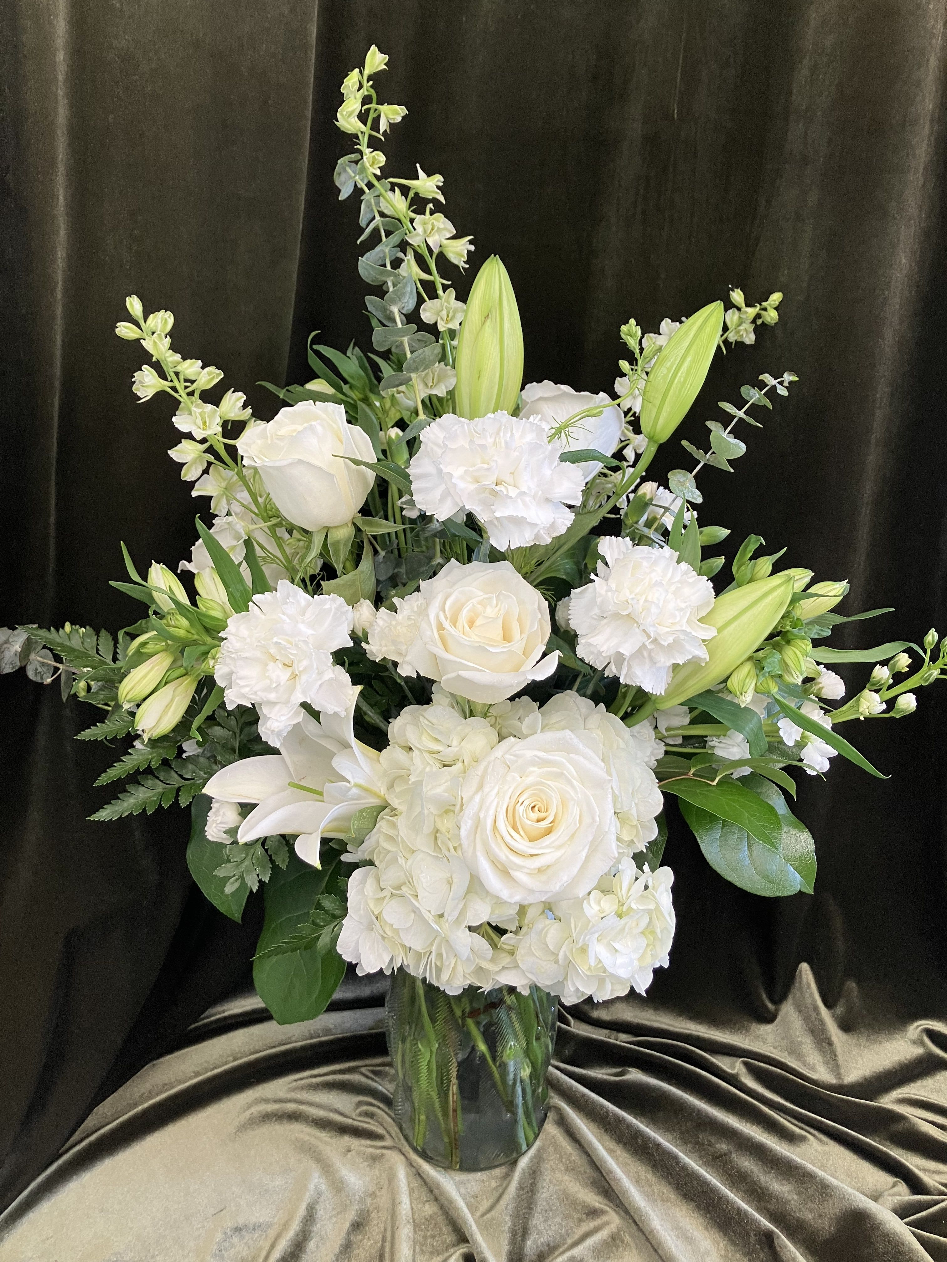 Traditional All White Arrangement - A classic and serene Arrangement, this flower arrangement is perfect as a sympathy bouquet, new baby, or a gentle "thinking of you" surprise. The white palette of roses, carnations, hydrangea bloom and lily buds is softened by green accents and branching stems, creating a formal yet approachable silhouette that makes the recipient feel comforted and seen.  Arranged in a clear glass vase, the design is layered and upright: compact hydrangea clusters add body, roses and carnations offer timeless texture, and the lily buds and snapdragon-like accents give vertical interest and understated movement. The piece is generous without being fussy, suitable for a mantel, bedside table, or reception desk.