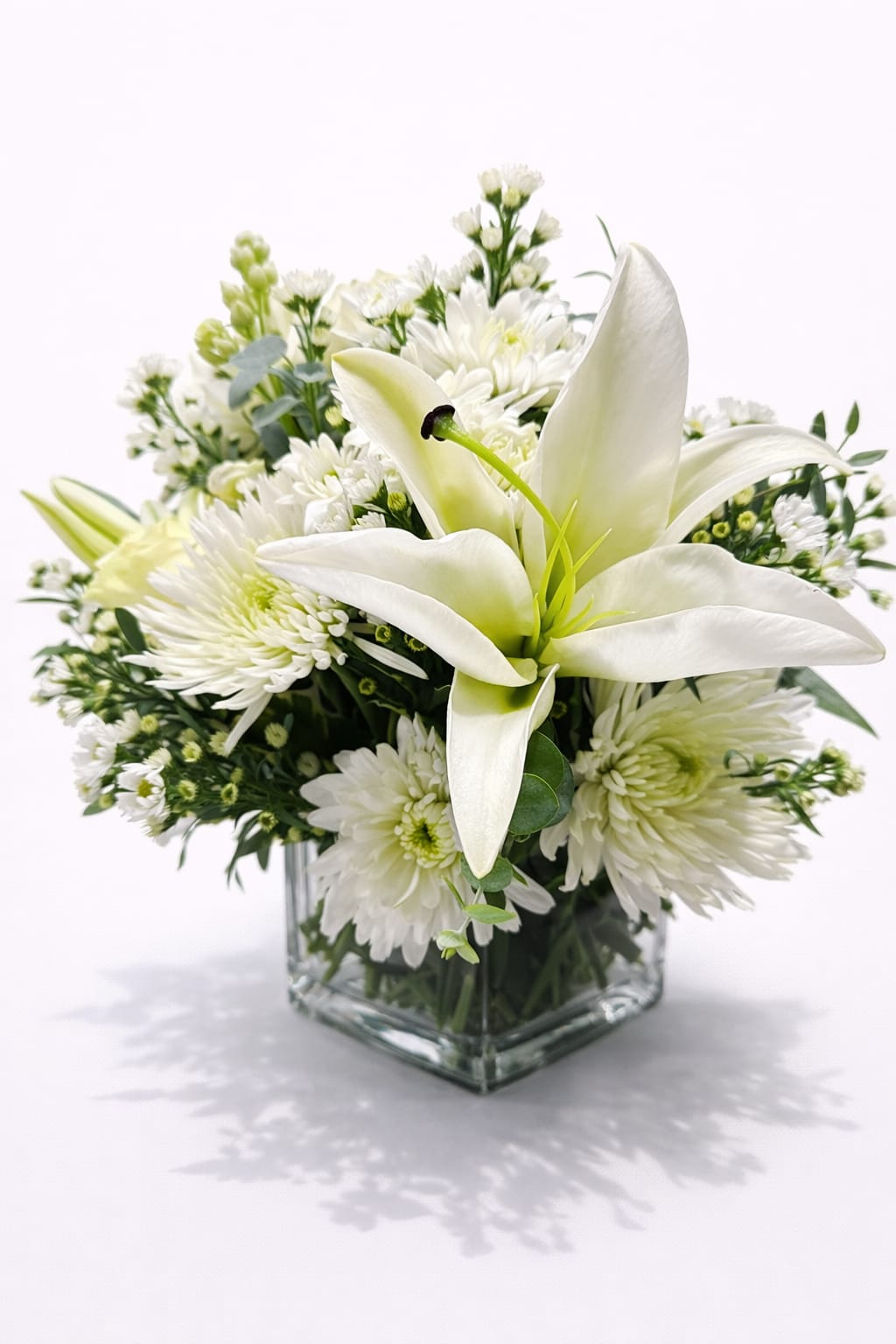 Lily Bliss White Lily and Chrysanthemum Arrangement - White lilies rise above a rounded cluster of white chrysanthemums and small daisy-like blooms in this Lily Bliss white lily and chrysanthemum arrangement-an elegant choice for a housewarming gift, a wedding centerpiece, or a thoughtful sympathy or "I'm sorry" gesture. The all-white palette with soft green accents and touches of pale yellow creates a serene, comforting impression that says "thinking of you" or "get well soon" without overwhelming the moment.  This compact design is tightly gathered in your choice of a clear glass cube or clear glass vase so stems and foliage show through for a clean, modern look. Alongside lilies and chrysanthemums you'll find daisies and rose accents that add texture and gentle contrast, with snapdragon buds and greenery woven in for height and movement. The arrangement reads as calm and composed-about the scale of a rounded centerpiece-making it easy to place on a bedside table, mantel, or entry console.  Give this bouquet to someone who needs a quiet, sincere lift: it comforts at a funeral or sympathy visit, celebrates a new home, or offers a graceful "thank you" or apology. Designed to feel fresh and long-lasting, keep blooms in cool, clean water and out of direct heat for best results; Flowers for Ladies &amp; Events delivers to Newington, CT for prompt local presentation.