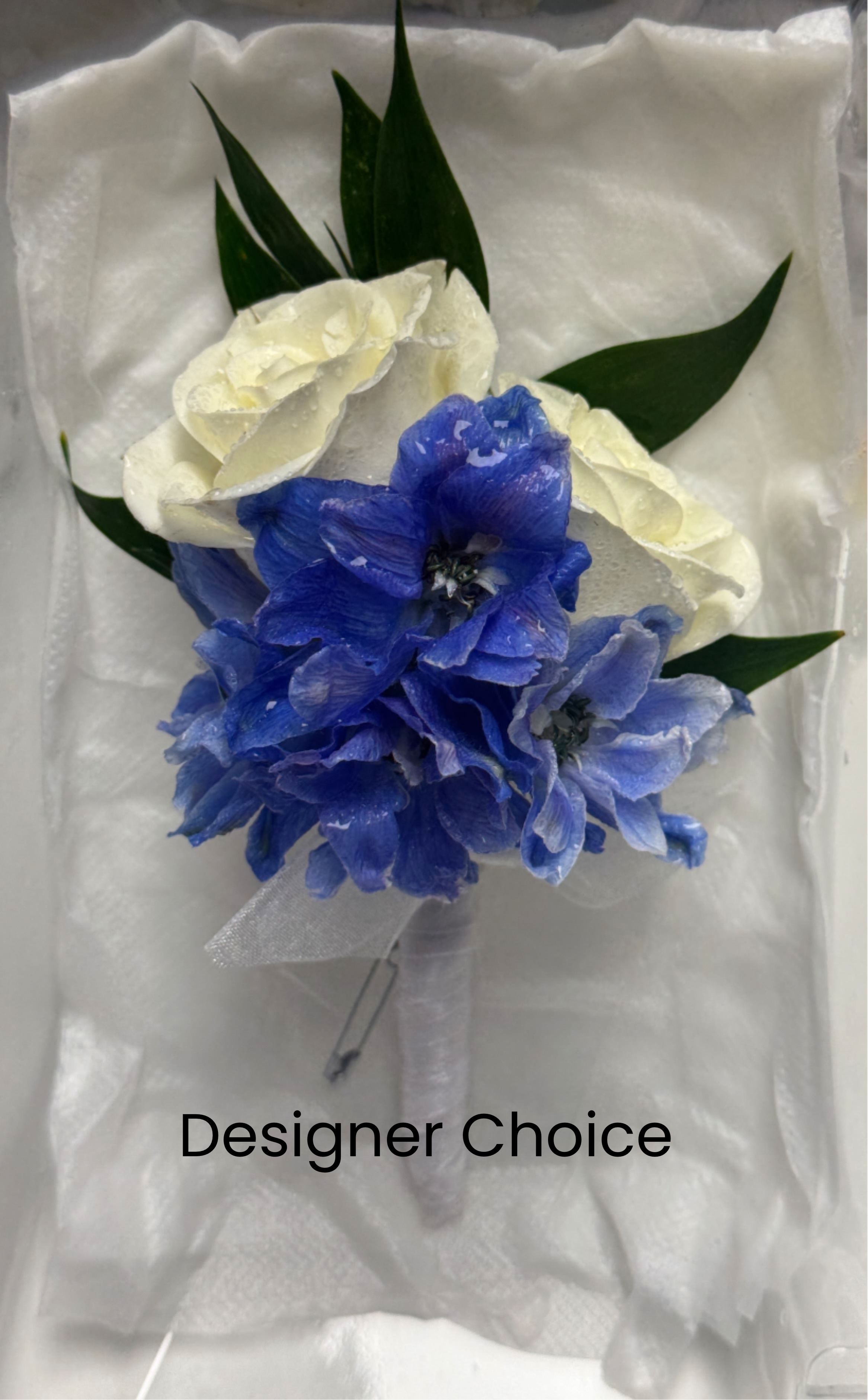 White rose and blue delphinium boutonniere with green leaves on white padding