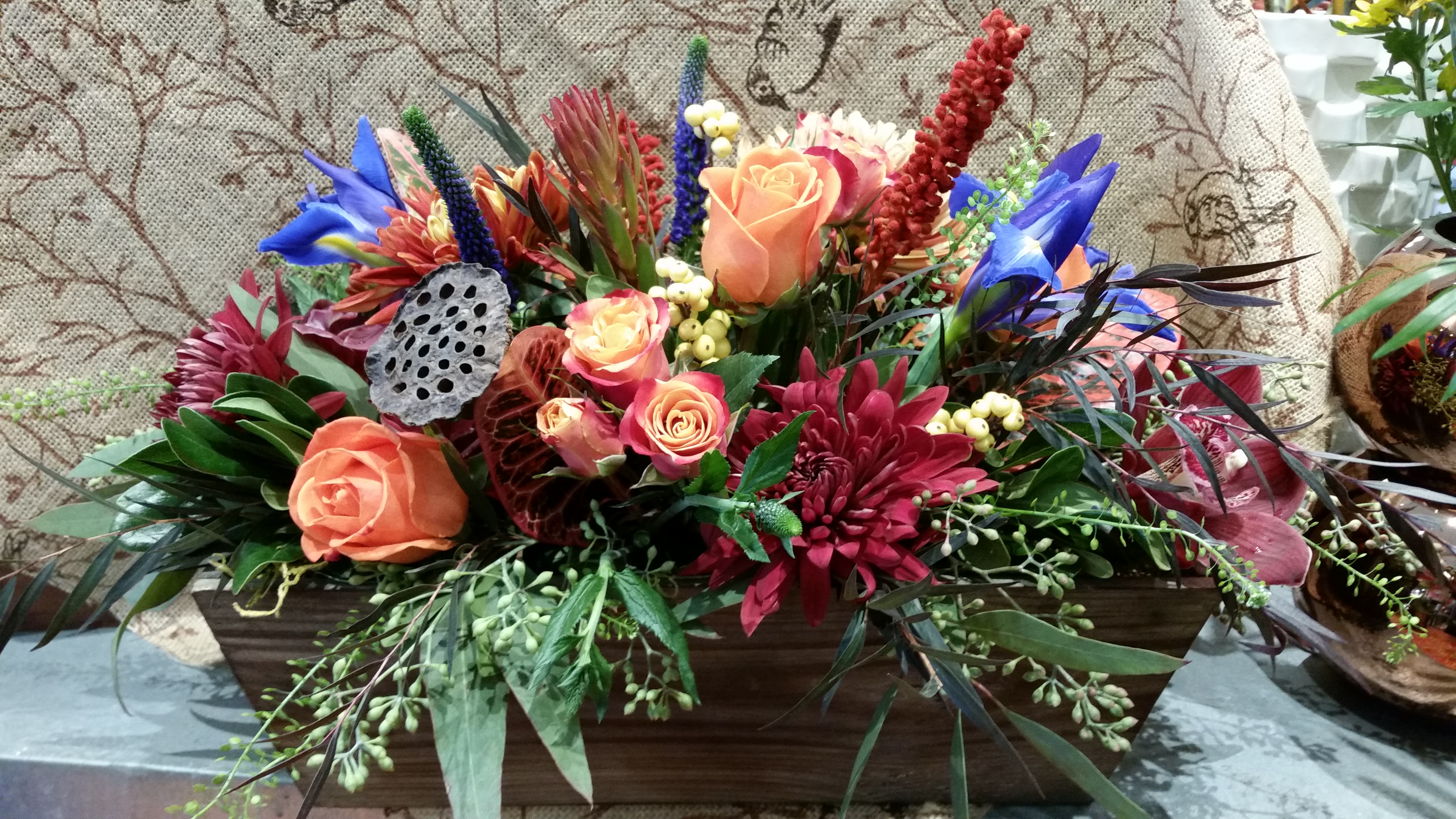 Barnwood Box Rustic Centerpiece - It starts with a simple wooden box.  Inside this box, we've combined a wonderful mix of garden blooms that will make your guests feel right at home.  Nothing pretentious here!  Roses, Iris, Chysanthemum, Orchid, and two varieties of eucalyptus create an interesting textural and colorful arrangement.  We just love the simple elegance of this piece.