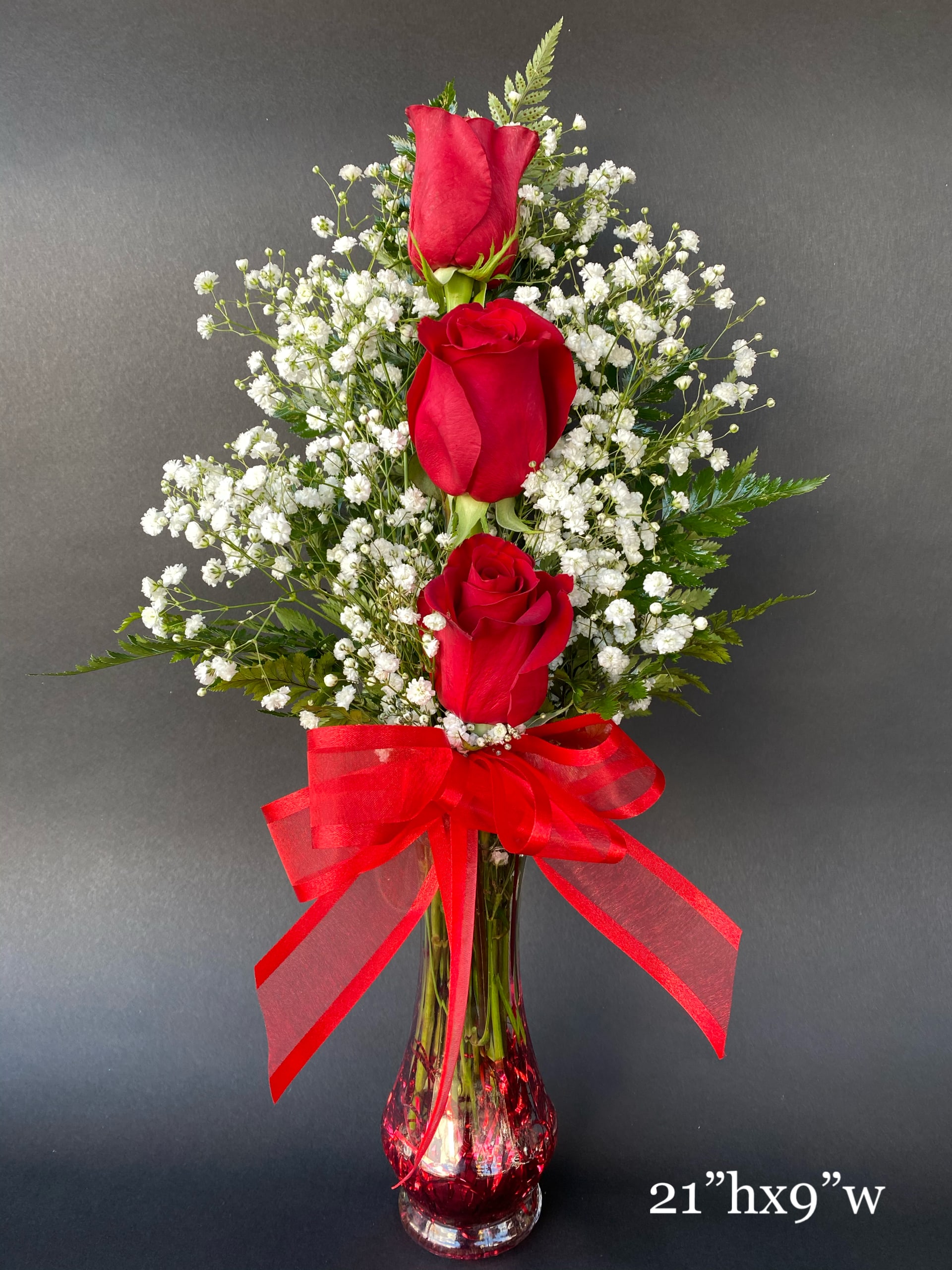 Three red roses in a clear vase with baby's breath and a red ribbon