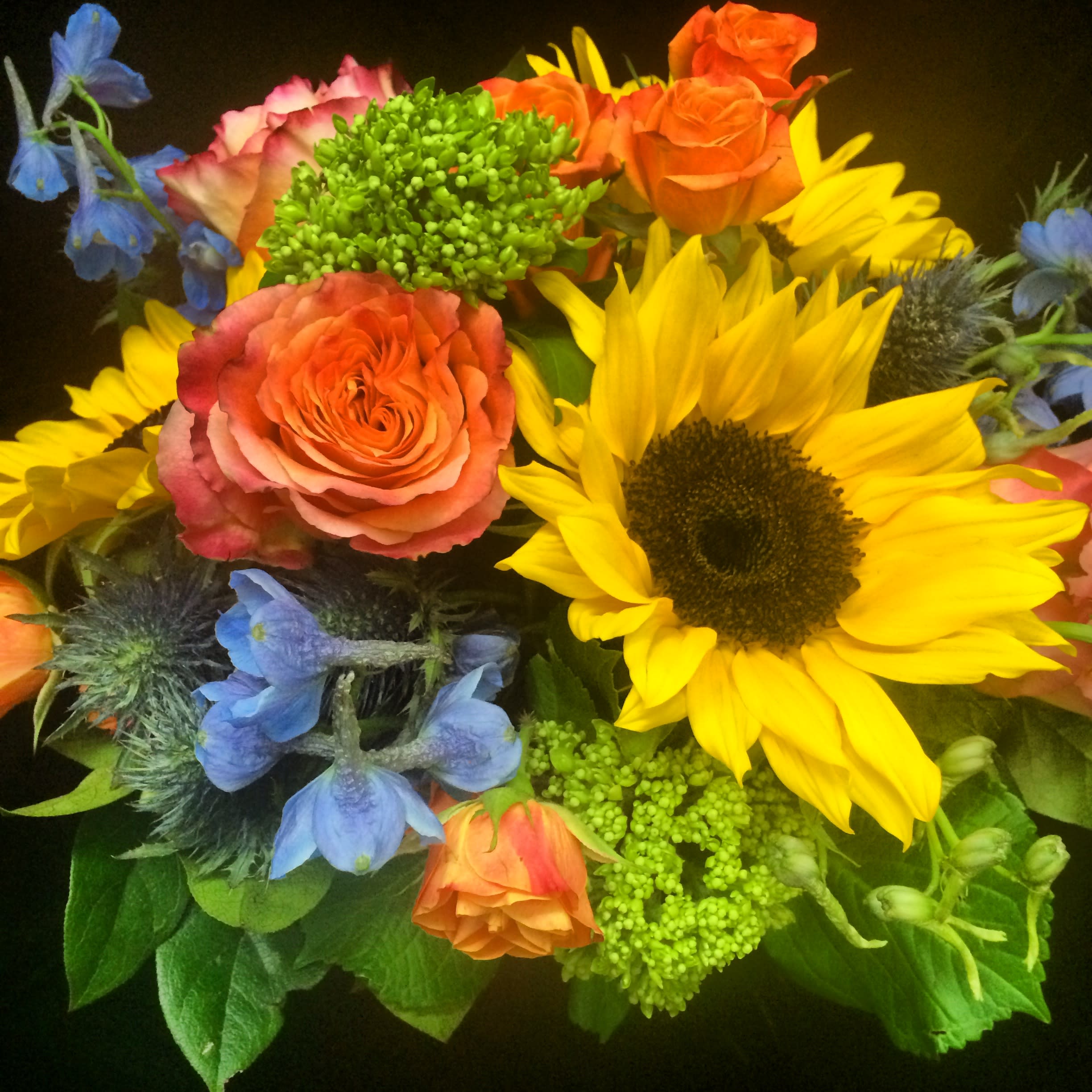 Bright bouquet with yellow sunflower, orange roses, blue delphinium, and green accent blooms