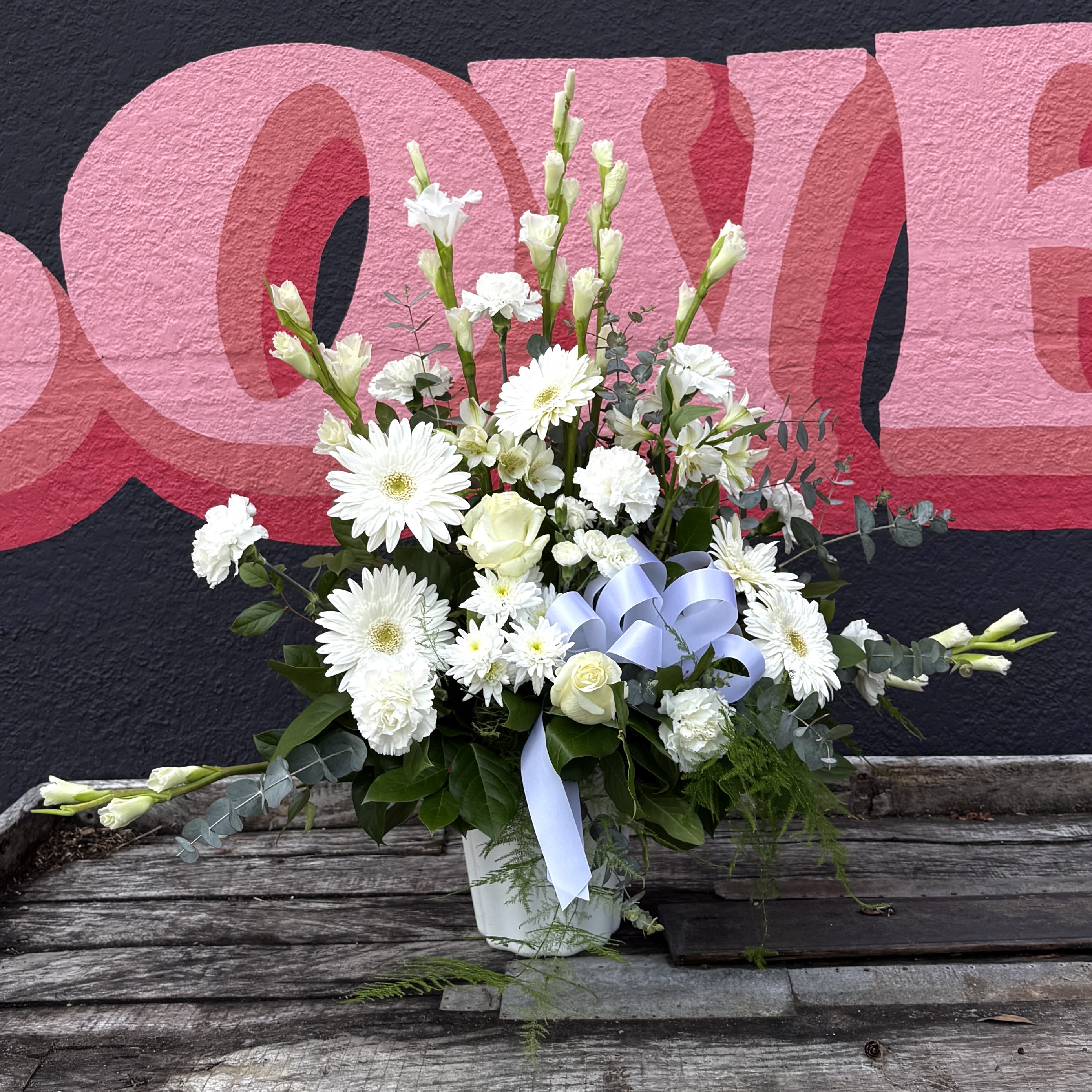 Floral Mache - A serene fan-style sympathy flower arrangement designed to bring quiet comfort and graceful presence, ideal as a funeral or memorial tribute. This floral arrangement for funerals features predominantly white blooms layered in an asymmetrical, vertical fan that reads calm and respectful-accented by a pale blue ribbon bow tied on a white ceramic container. As a Baltimore florist offering compassionate flower delivery in Baltimore, this design is perfect for honoring a life with understated elegance.  The arrangement emphasizes white and pale tones for a soothing, peaceful effect, with subtle touches of pale blue from the ribbon and gentle green foliage for balance. The piece sits in a white ceramic container and measures to suit typical memorial spaces-please allow 24-48 hours notice for preparation.  For those seeking a dignified floral expression from a trusted local florist, this arrangement conveys sympathy and support with quiet beauty. The Flower Cart offers reliable same-day and scheduled options for flower delivery in Baltimore to funeral homes, churches, and residences-contact us for details and to confirm availability.