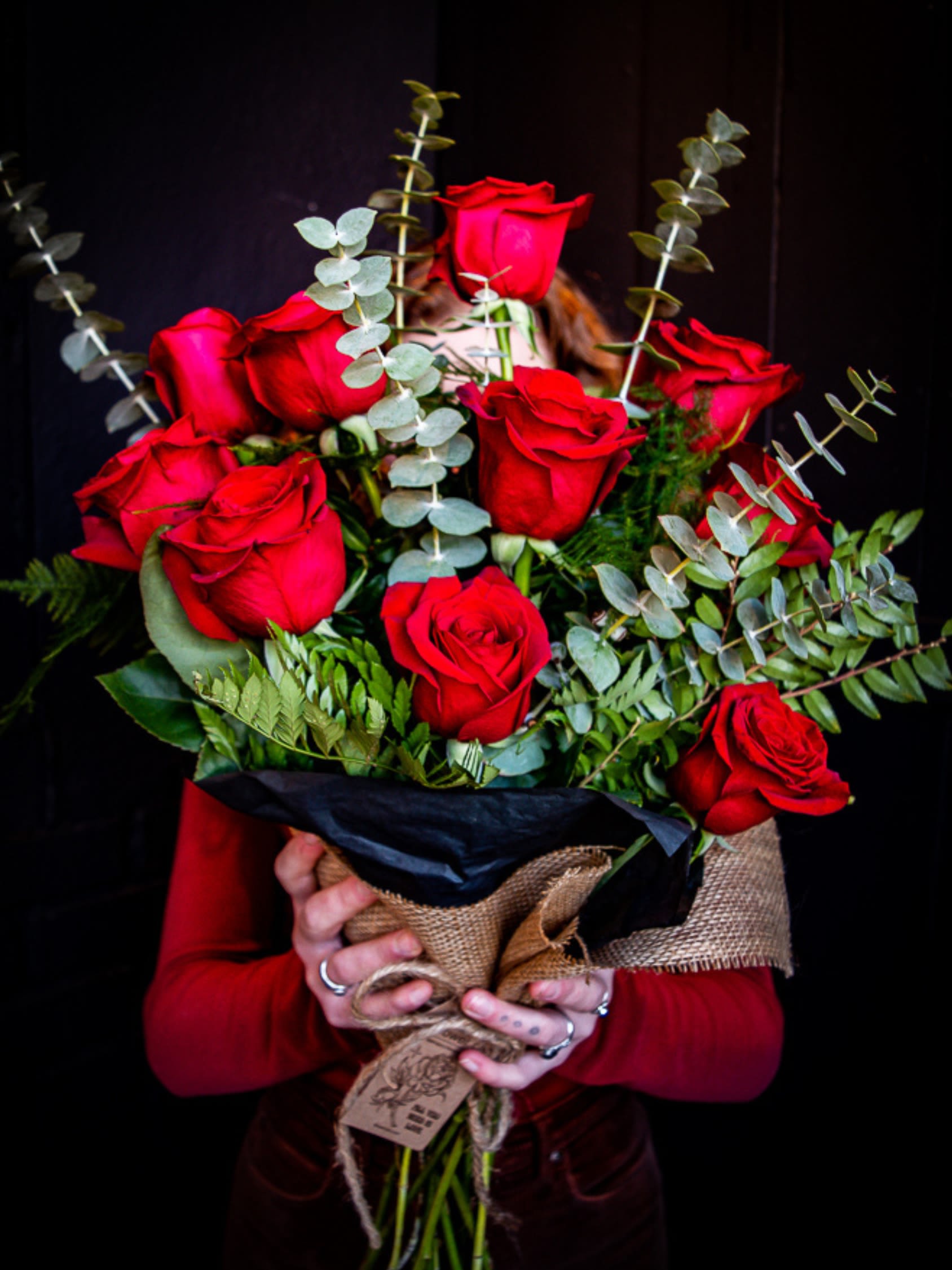 Hand-tied bouquet of red roses wrapped in burlap and black paper, held against a dark background