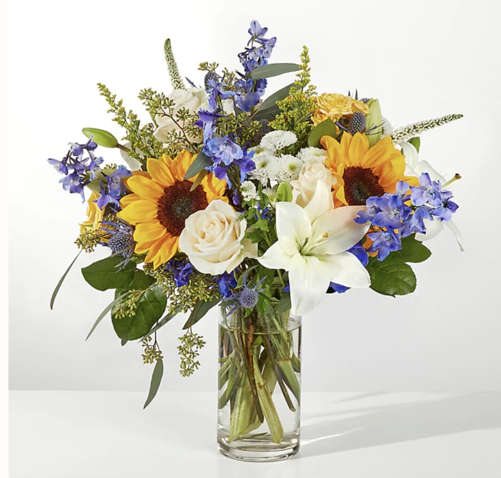 Bouquet of sunflowers, white lilies, and blue flowers in a clear glass vase