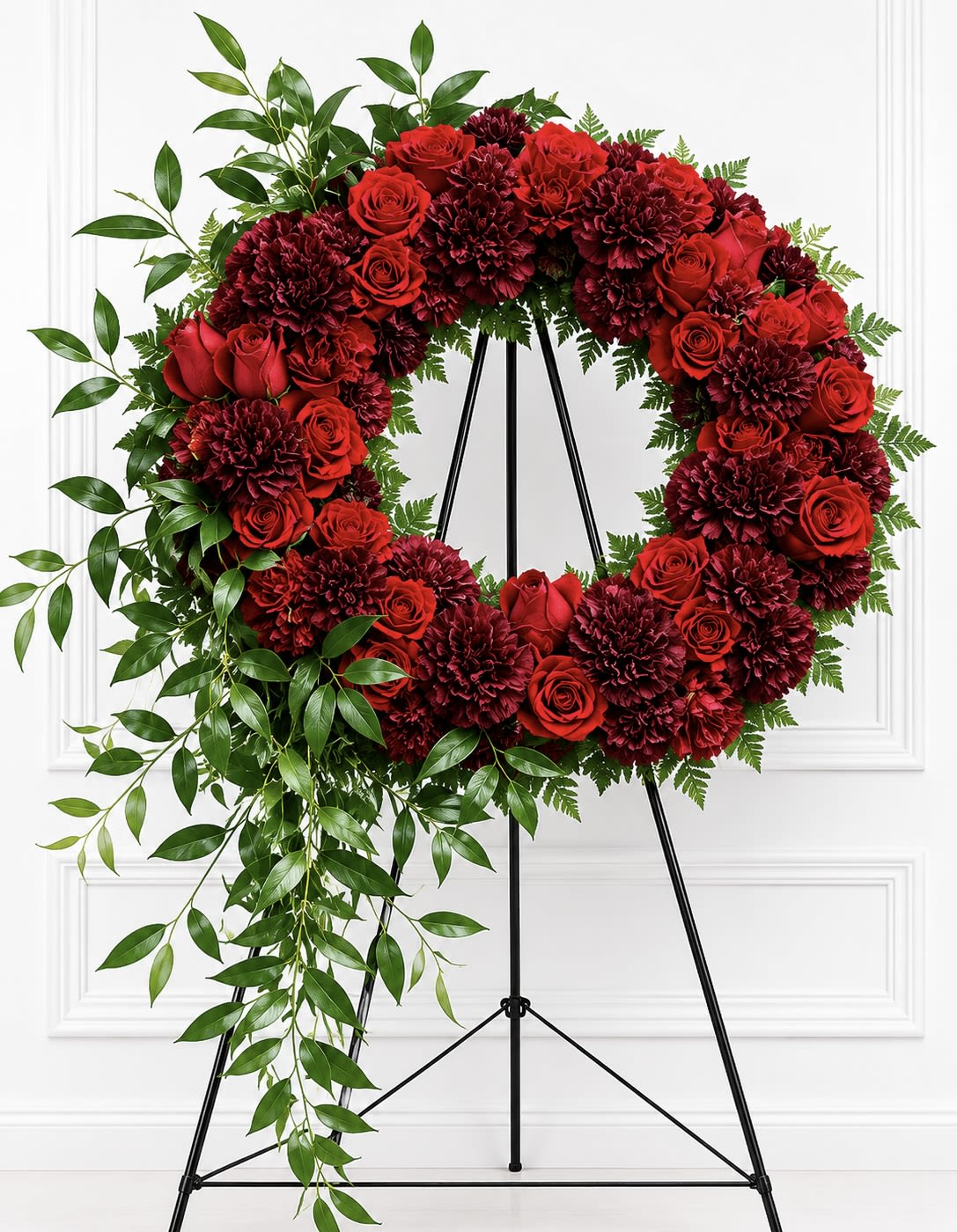Crimson Red Sympathy Wreath - Deep crimson roses and deep burgundy carnations form a large, circular sympathy wreath that reads as both solemn and elegant-the kind of funeral arrangement that helps the person you're remembering feel honored and truly seen. The rich red and purple tones against lush green trailing foliage create a dramatic, formal statement suited for a funeral service or memorial setting.  Dense layers of roses and carnations create a full floral ring, with a cascade of greenery flowing down the left side for graceful movement and contrast. The wreath is displayed on a black metal easel stand for a traditional, ceremonial presentation; white blooms subtly punctuate the design to add gentle highlights and depth. This wreath is sized and styled for placement beside a casket or at a memorial site-order with confidence for sympathy services year-round.
