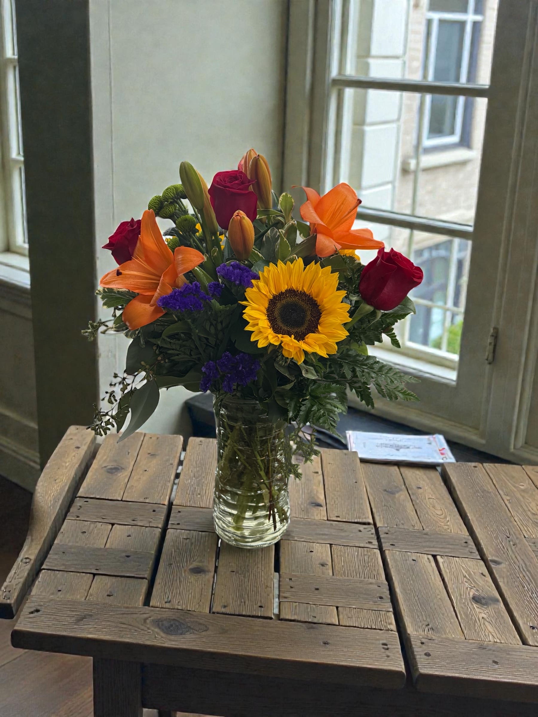 Bouquet of sunflowers, red roses, orange lilies, and purple filler in a glass vase