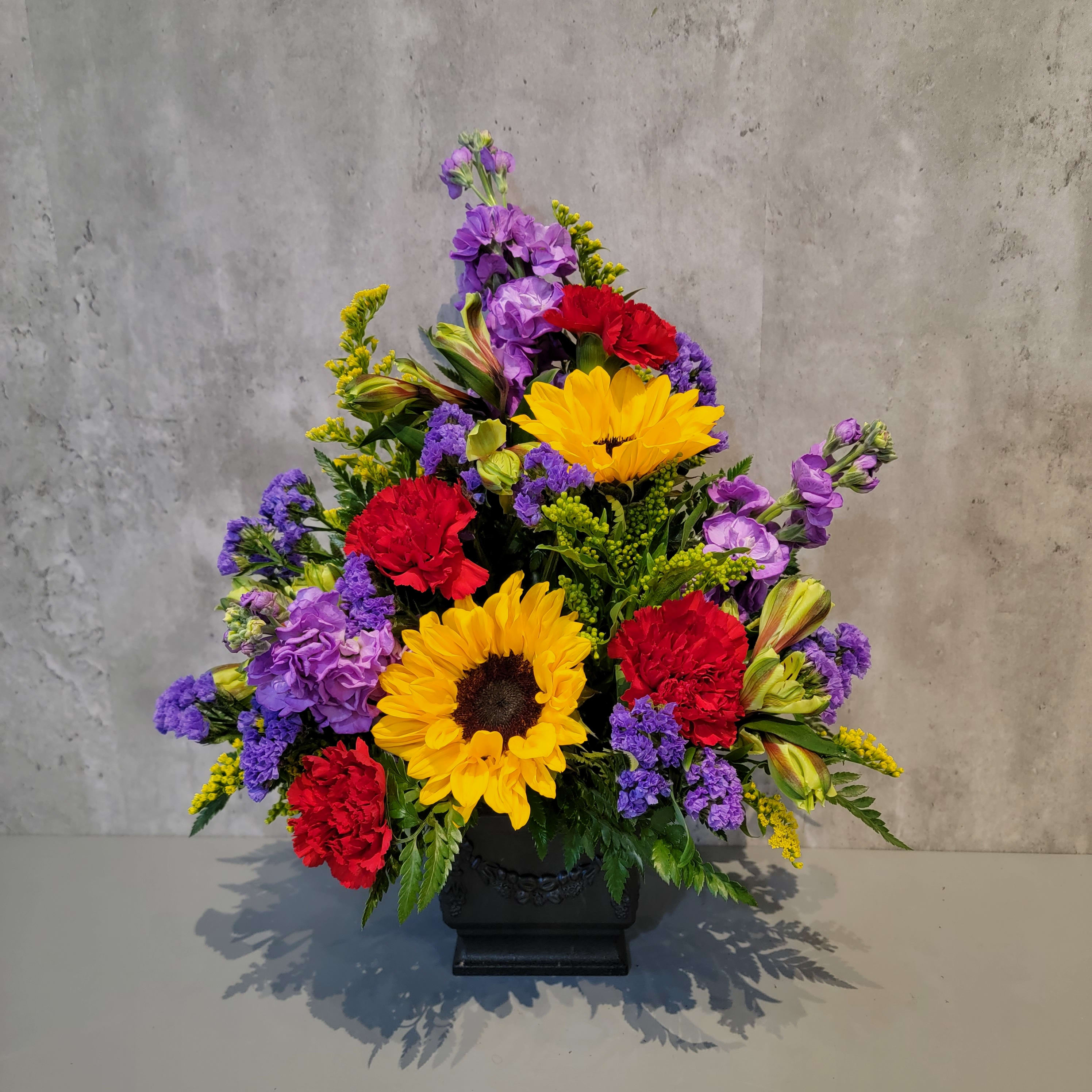 Colorful floral arrangement with sunflowers, red carnations, and purple blooms in a black vase