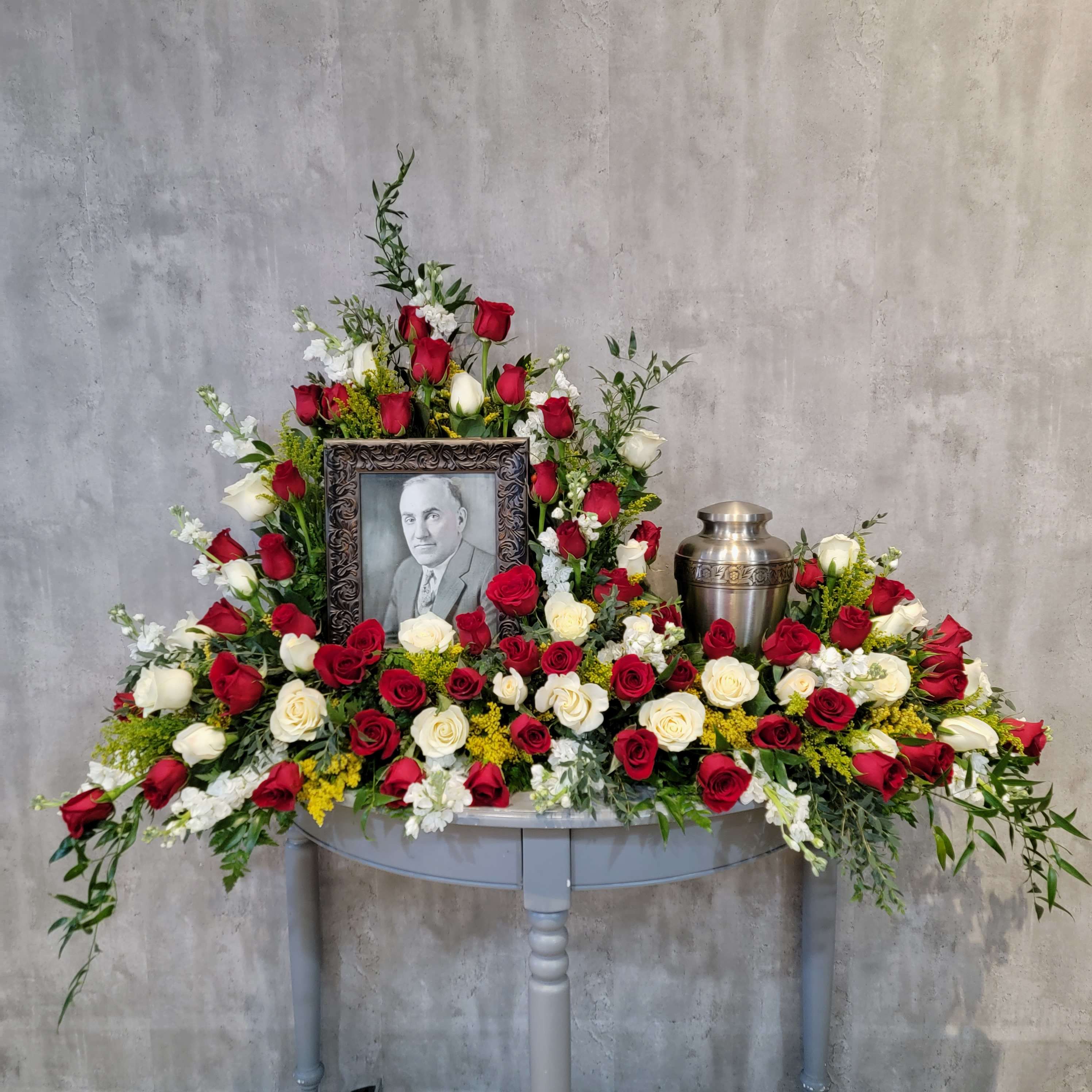 Sympathy floral arrangement with red and white roses around a framed portrait and urn