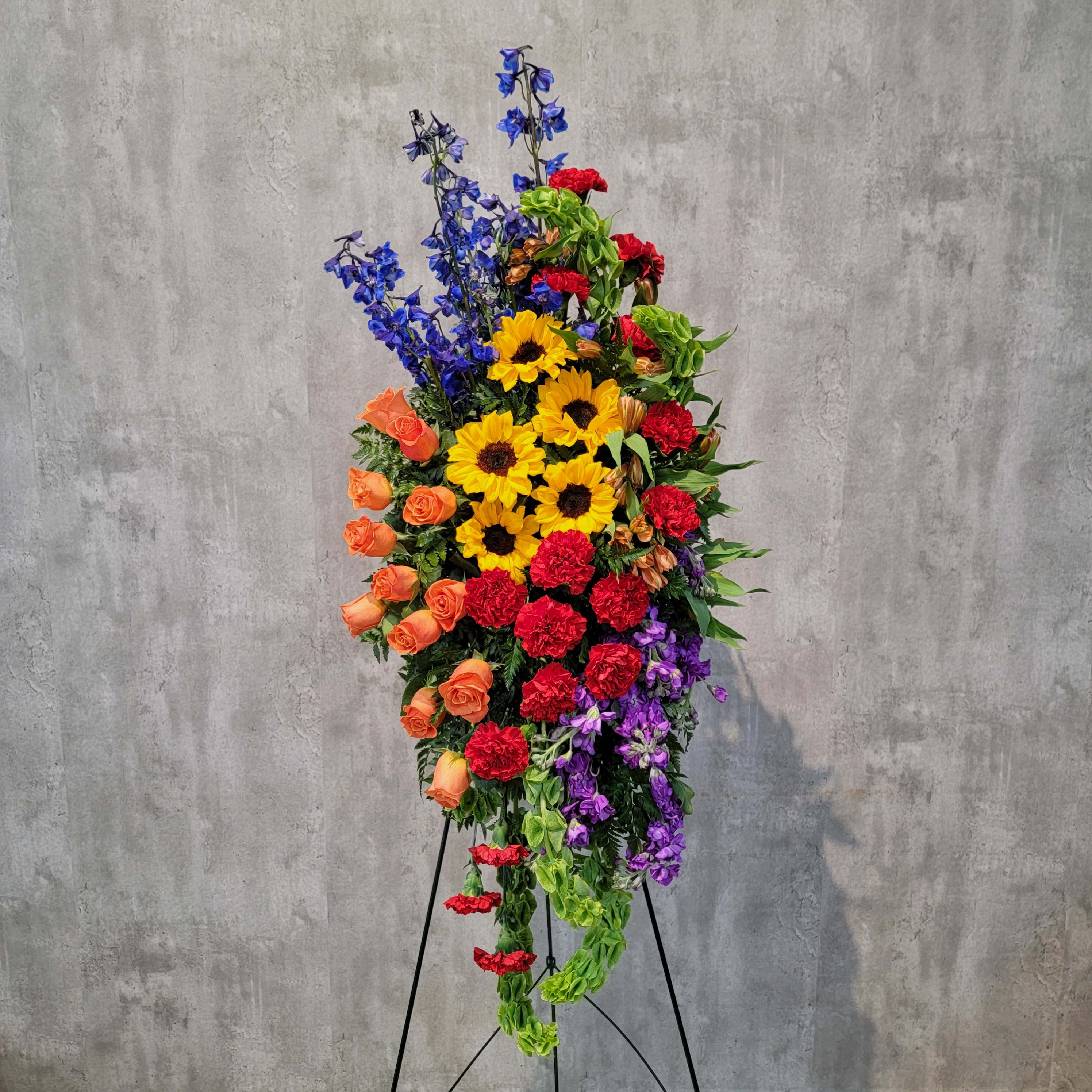 Tall standing floral spray with sunflowers, roses, carnations, and blue delphinium