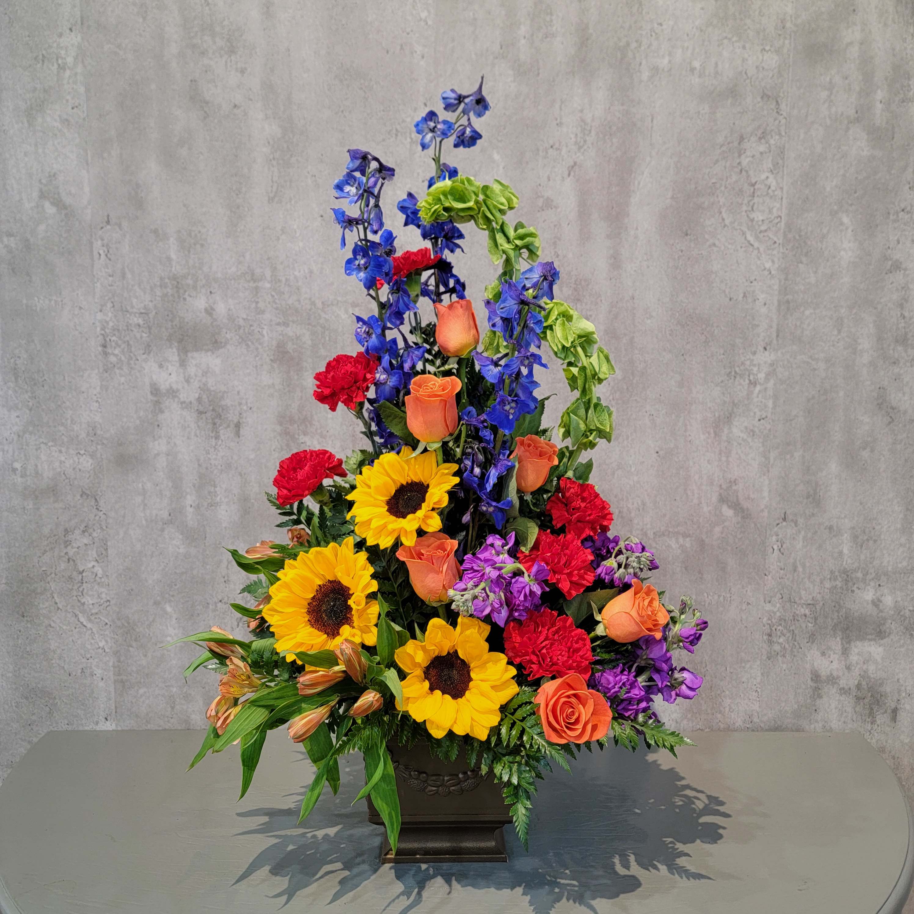 Tall mixed flower arrangement with sunflowers, roses, and blue delphinium in a dark vase