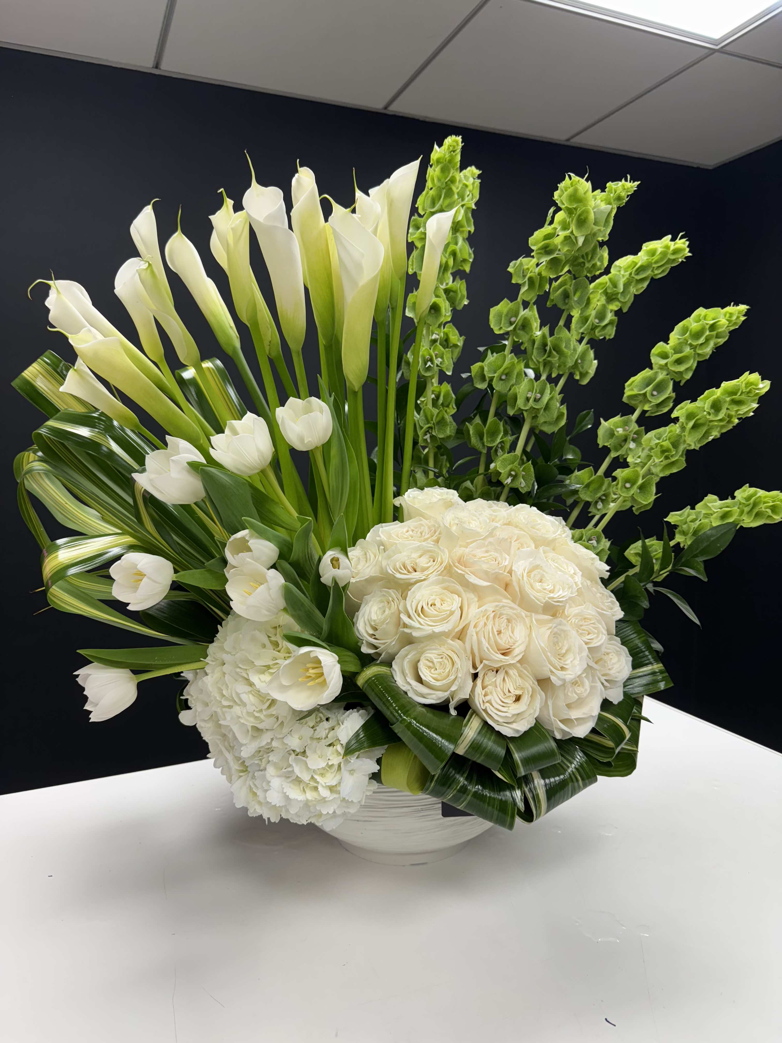 Tall white arrangement of calla lilies, roses, tulips and hydrangea in a low white bowl with green accents