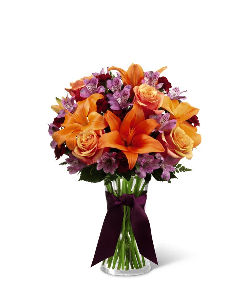 Vase arrangement of orange lilies, peach roses, and purple flowers in a clear glass vase with dark ribbon