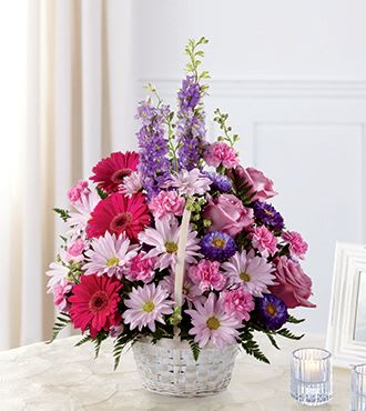 Pink and purple mixed flower basket arrangement in a white woven basket on a table