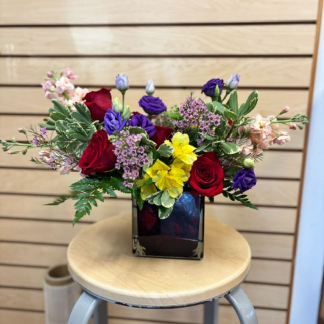 Mixed bouquet of red, purple, pink, and yellow flowers in a square vase