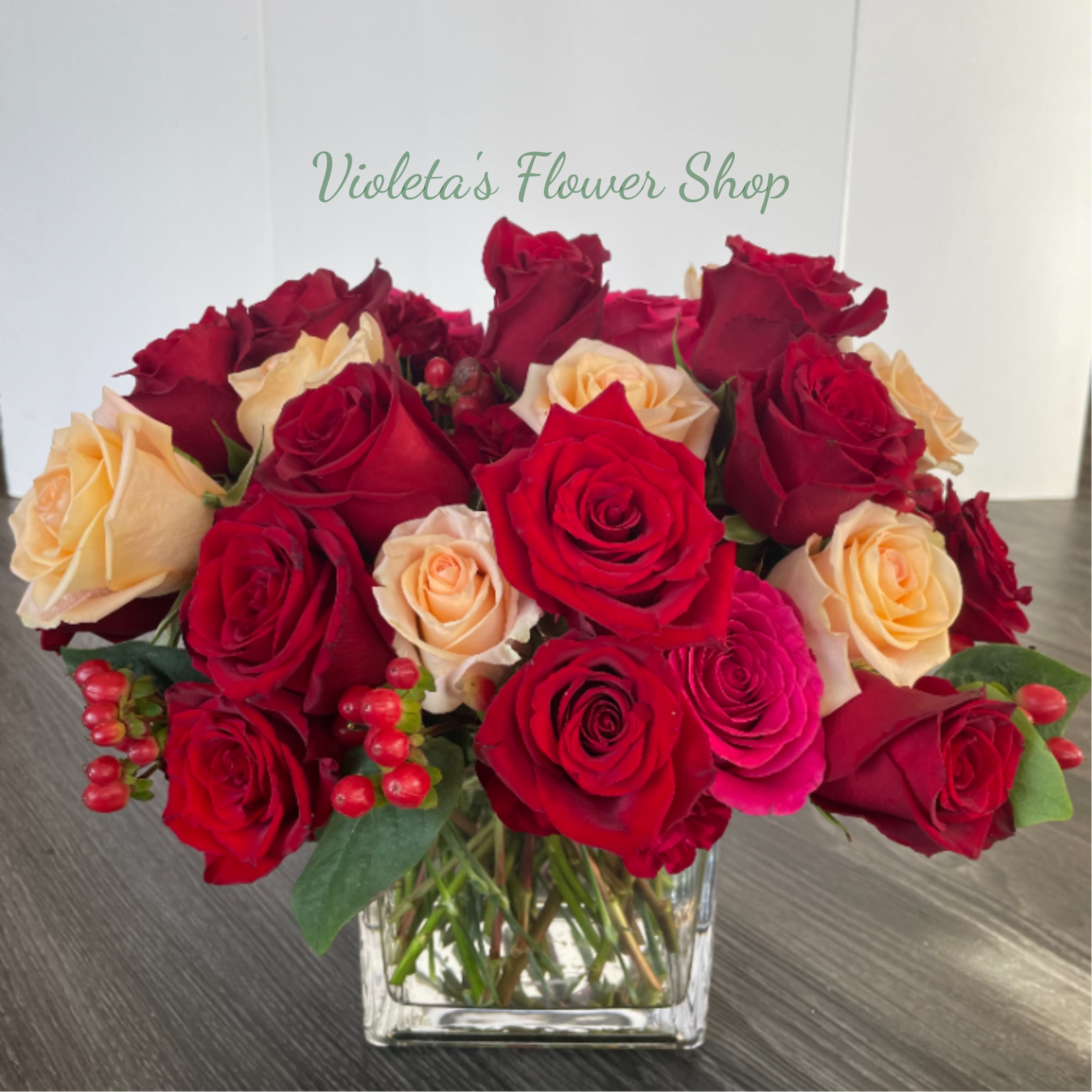 Totally In Love With You. - This arrangement uses only roses, densely packed to create a plush, painterly mix of saturated reds, lively pinks and warm peach tones. Small red berry accents tucked near the base add a hint of texture and festive sparkle, while the clear square glass vase gives the design a clean, modern anchor that suits both bedside tables and dining-room centerpieces. The low, rounded profile keeps the focus on the blooms' color and form, making it easy to place anywhere and enjoy up close.  Send this when you want the recipient to feel truly celebrated-an intimate anniversary gesture, a romantic note for love, or a sweet Thinking of You moment. 