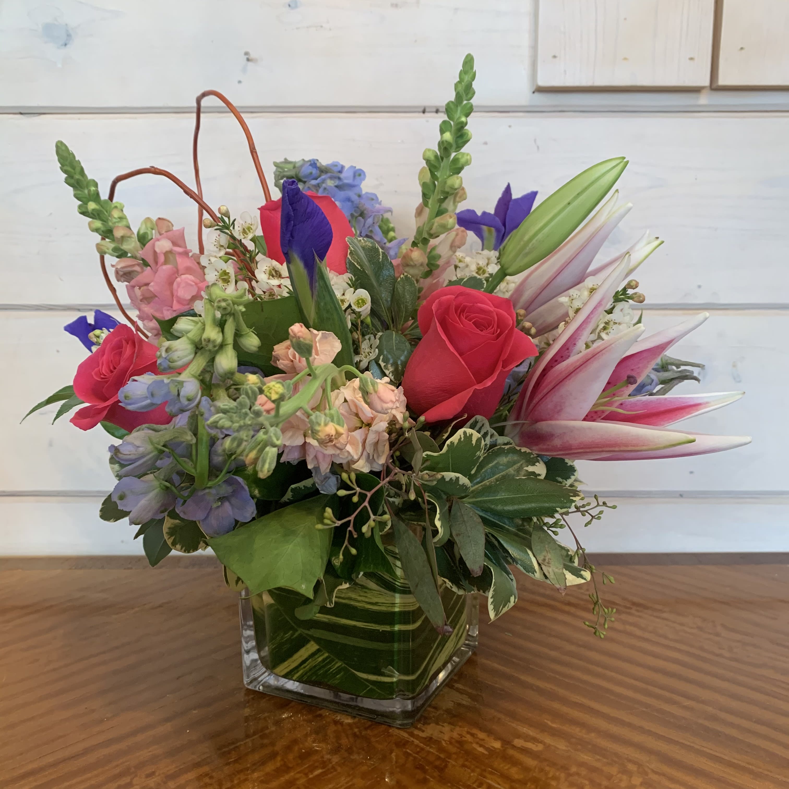 Mixed bouquet of pink roses, lilies, and blue flowers in a square glass vase