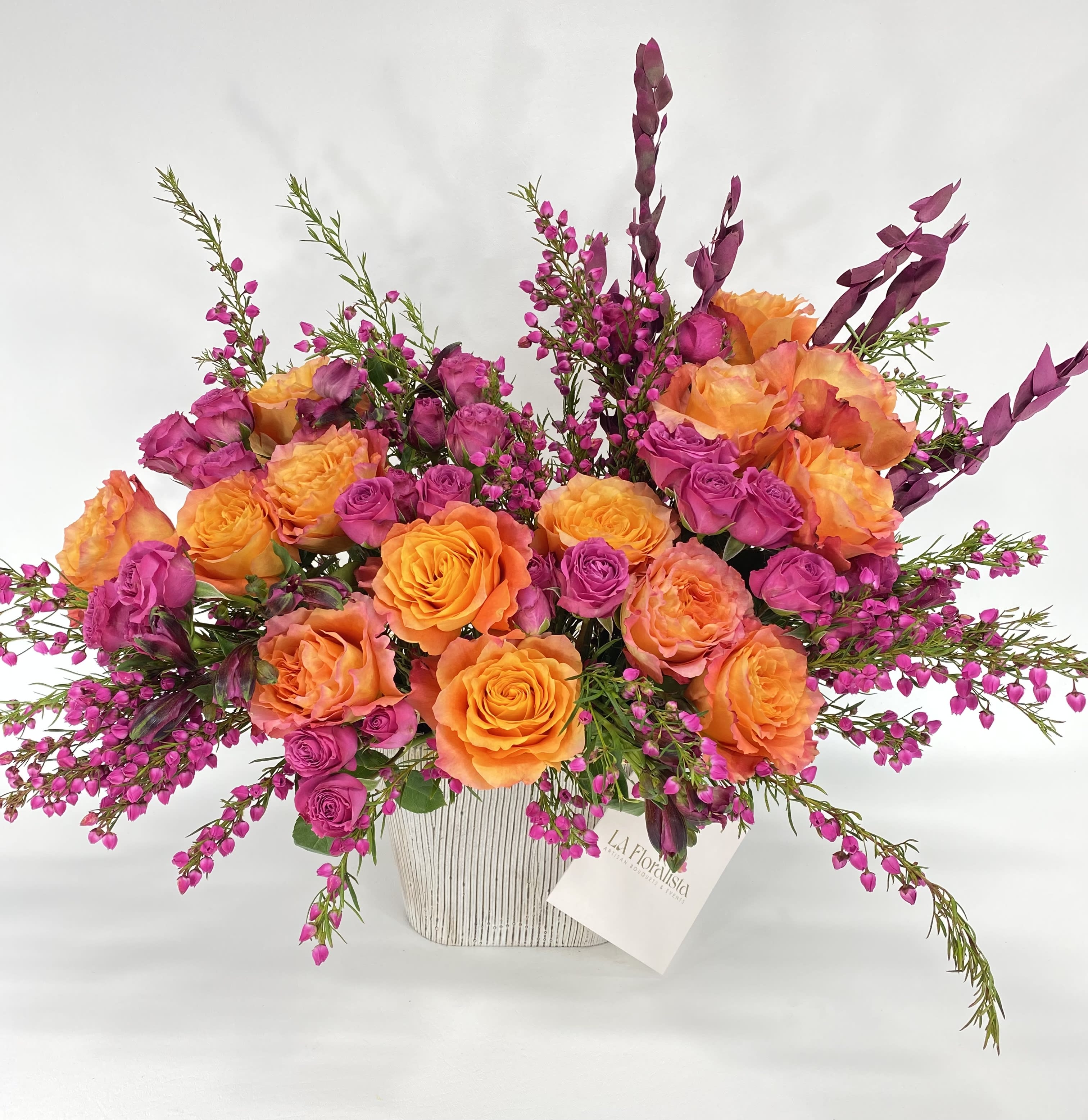 Orange and magenta roses in a white vase with purple filler flowers
