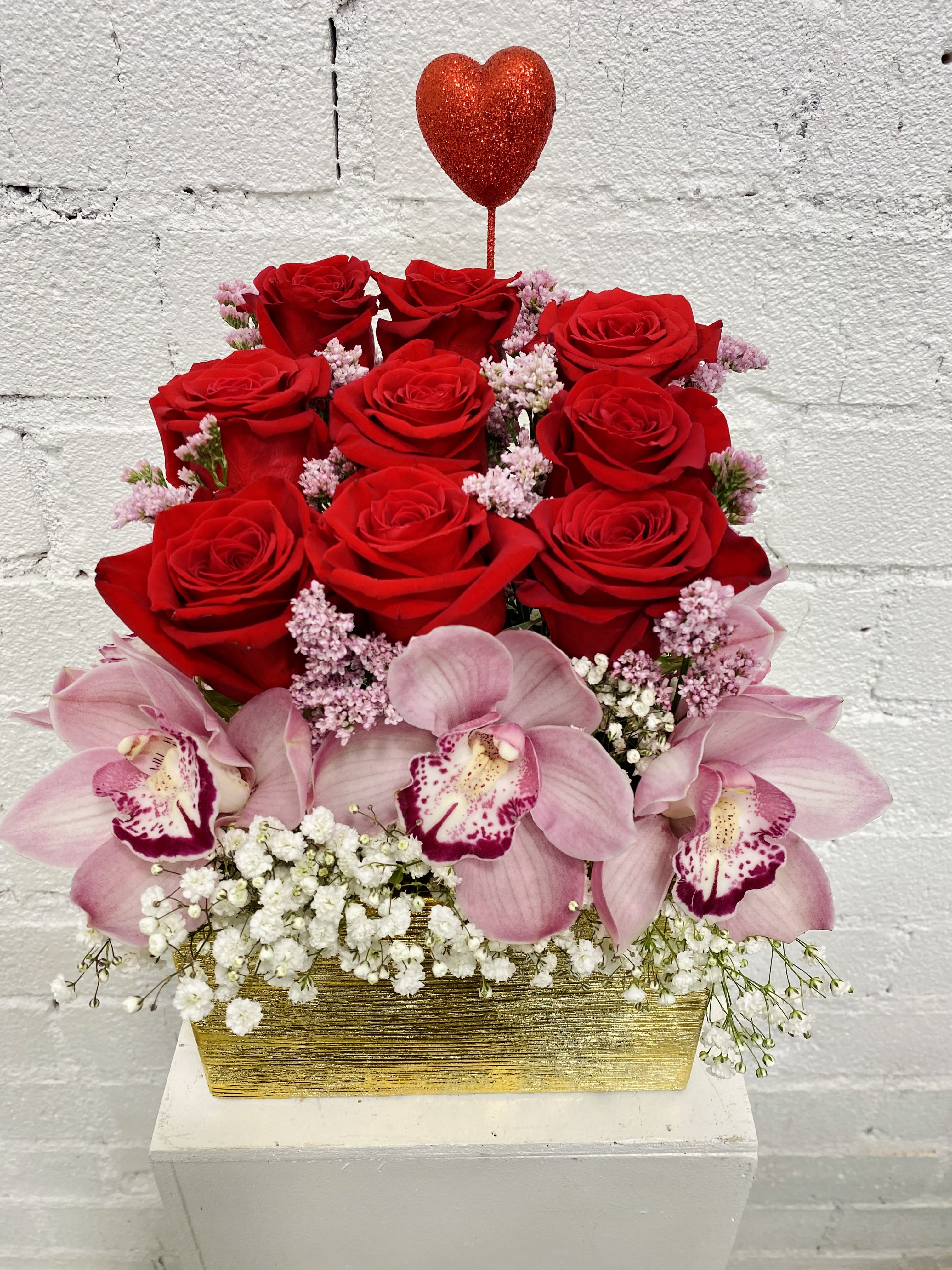 Red roses and pink orchids in a gold rectangular container with a heart pick