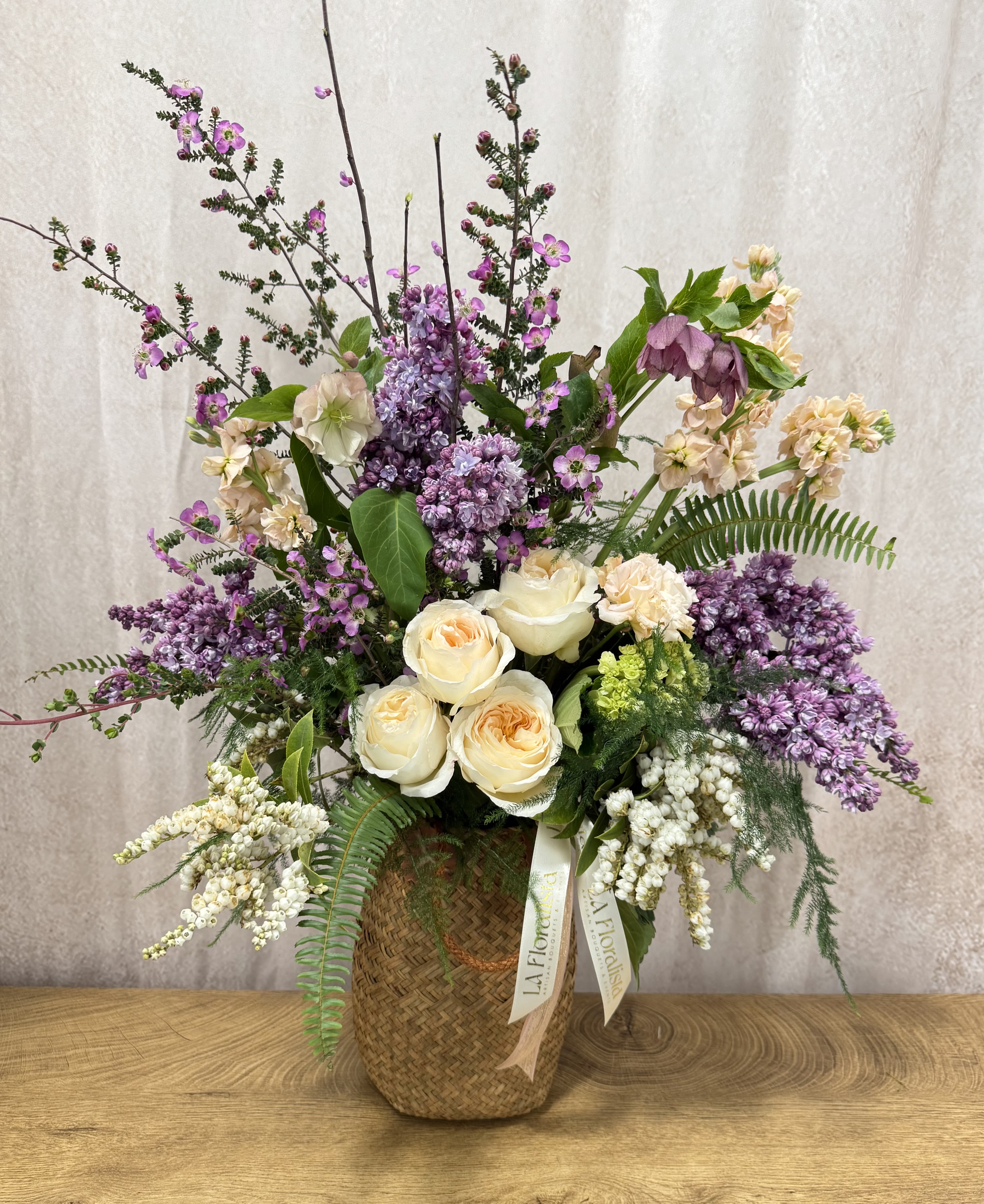 Tall floral arrangement with cream roses, purple blooms, and a woven basket vase