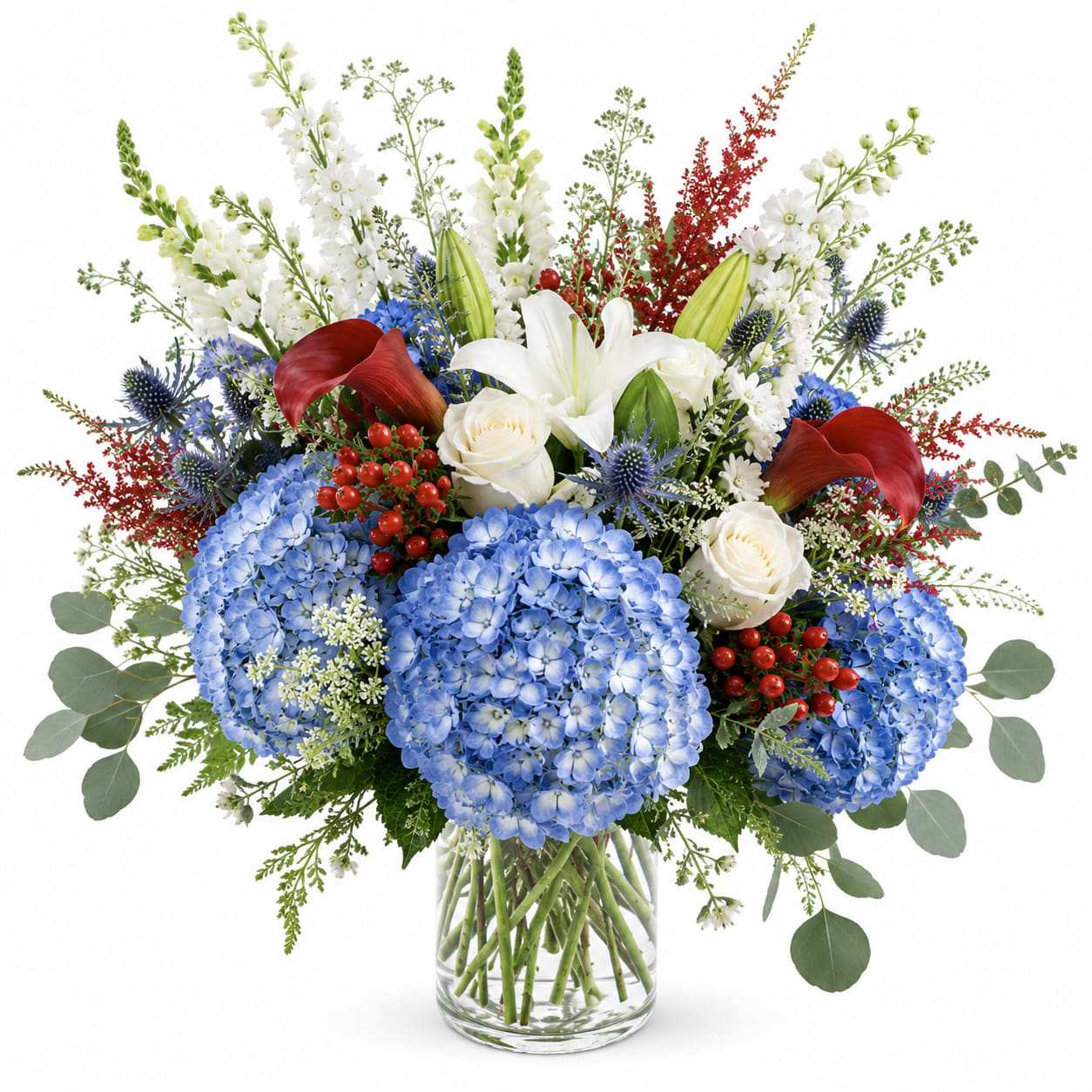 Patriotic Garden  - a lush, artistic arrangement inspired by elegant summer gardens and timeless patriotic hues. Soft blue hydrangeas are paired with creamy white roses,fragrant lilies,thistle,and a seasonal berries for a rich textured look . Designed in a clear glass vase with flowing greenery, this arrangement feels both refined and naturally romantic -perfect for celebrations, sympathy, gatherings, or an unforgettable statement piece.
