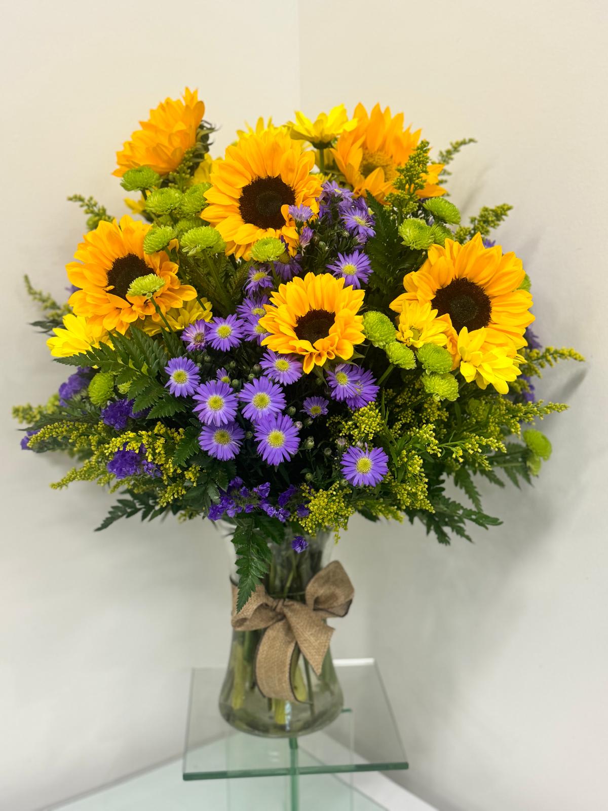 Tall arrangement of yellow sunflowers and purple asters in a clear vase with a burlap bow