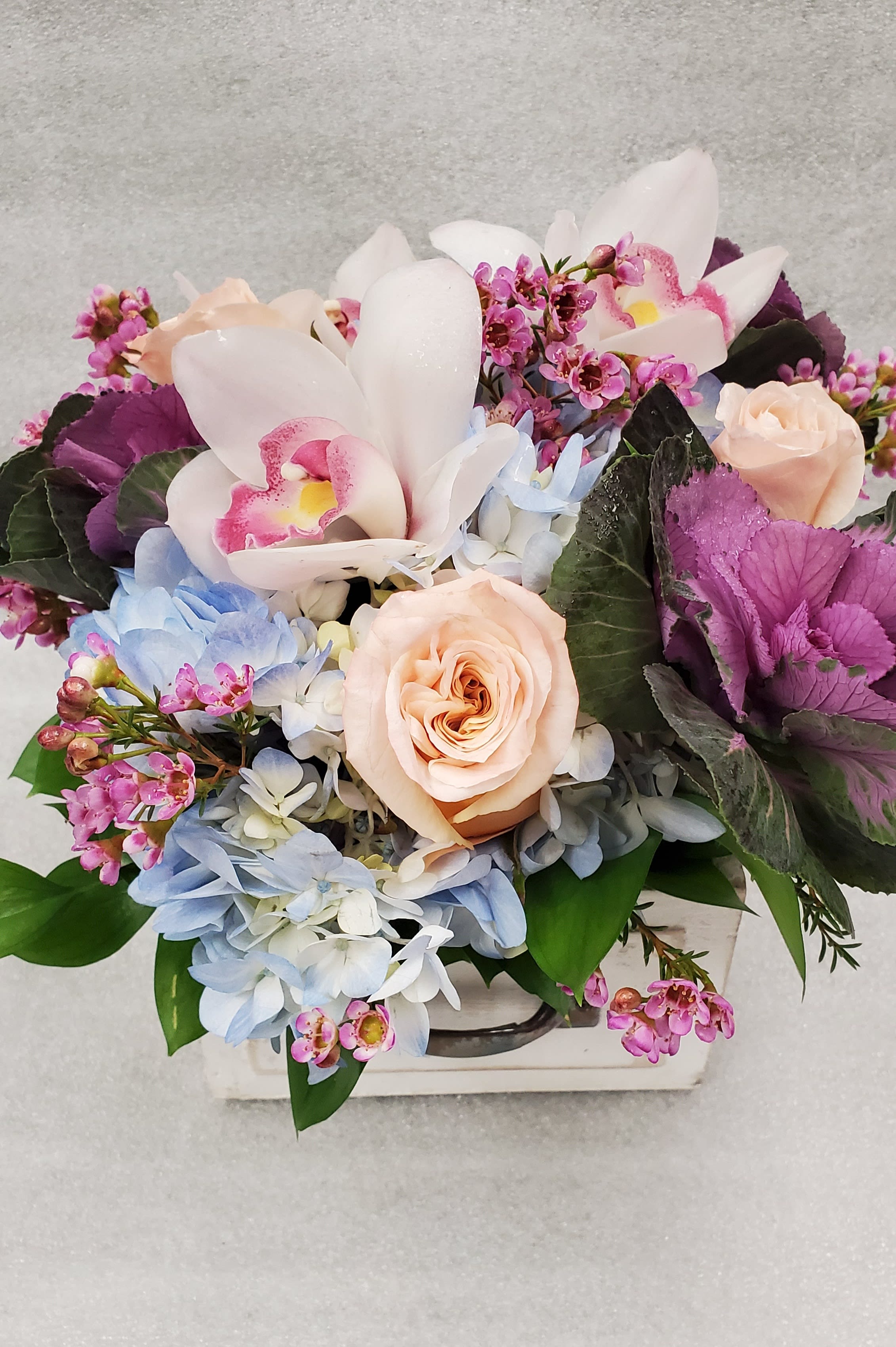 Mixed bouquet with pale roses, orchids, and blue hydrangeas in a white container