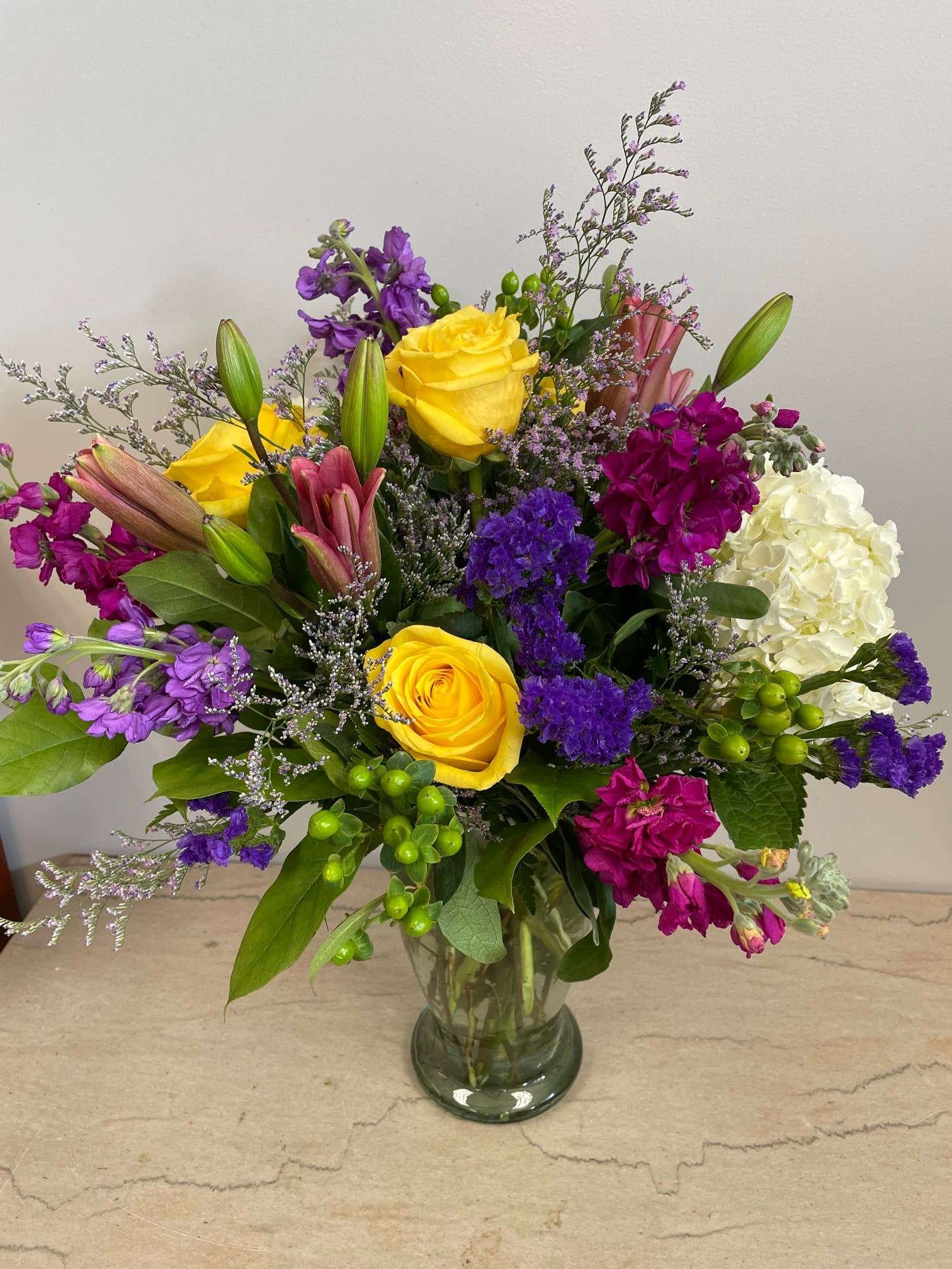 Mixed bouquet of yellow roses, purple flowers, and white hydrangea in a glass vase