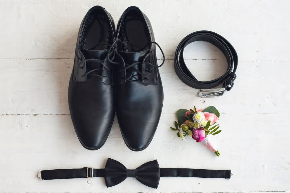 Black dress shoes, belt, bow tie, and a small boutonniere on a white surface