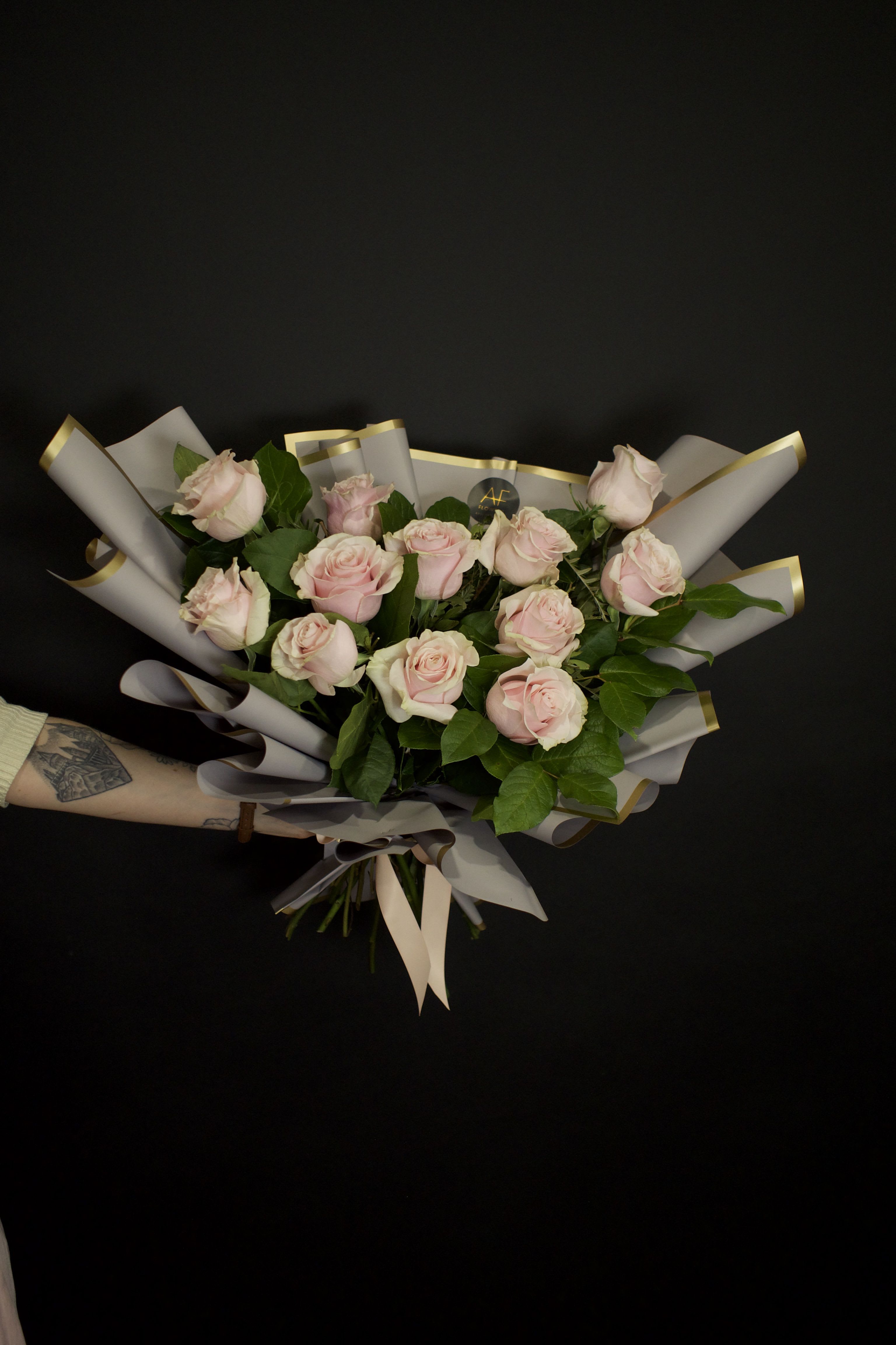 One dozen light pink roses - Hand tie bouquet - A contemporary bouquet of stunning light pink roses, thoughtfully designed with a modern aesthetic. Wrapped in sleek nude paper with elegant gold accents, this hand tied bouquet is also complemented by lush greenery. Finished with a luxurious ribbon, this bouquet embodies sophistication and timeless charm, making it the perfect gift for any stylish occasion.